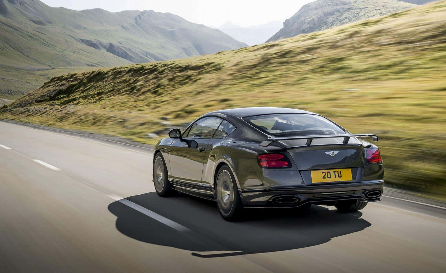 賓利Continental GT Sport 壁紙 — 一輛時尚的深綠色賓利 Continental GT 在明亮的日光下，沿著蜿蜒的山路疾馳，背景是模糊的草坡和遠處的山峰。