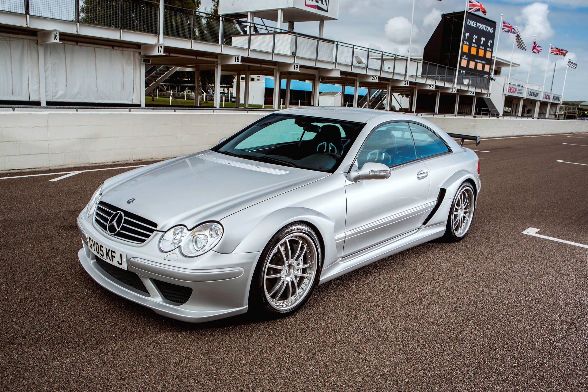銀色 Mercedes-Benz CLK GTR 賽車在賽道上，背景有看台和旗幟。