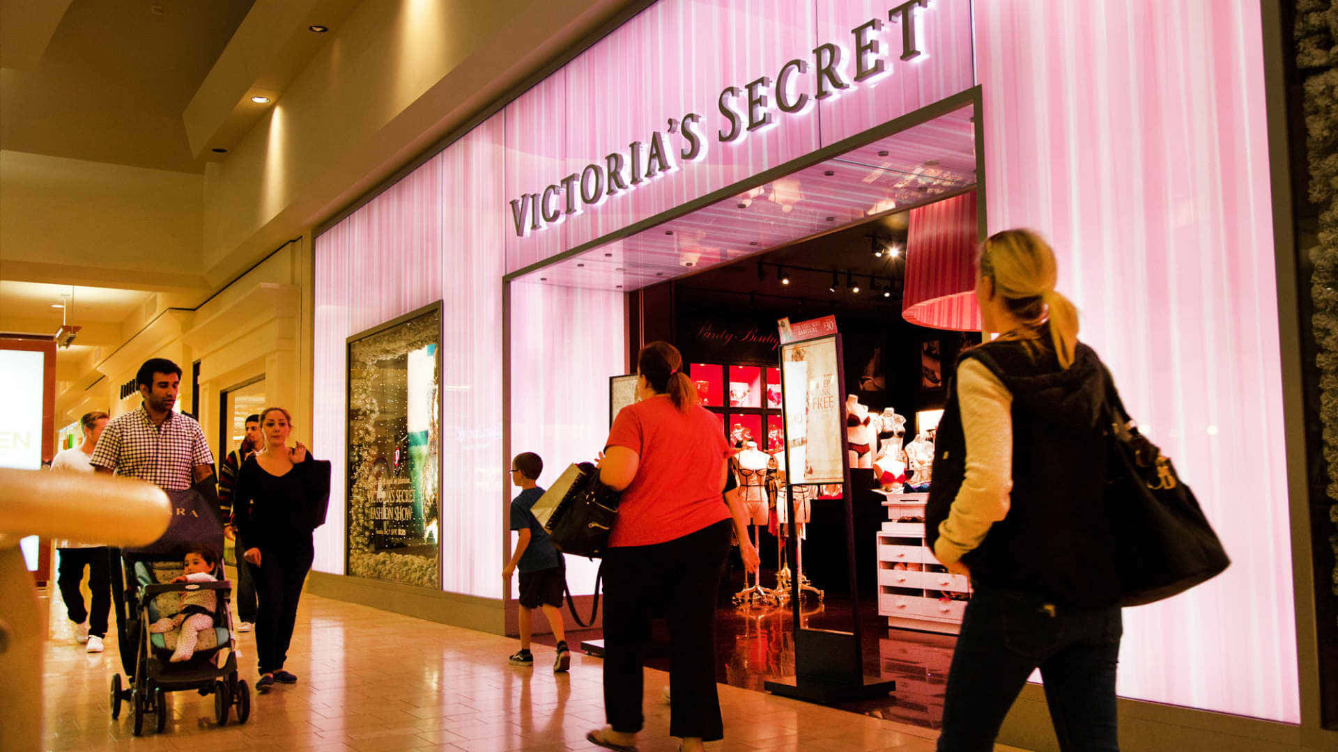 Victoria's Secret 桌布 壁紙 — 購物者漫步經過一間燈光明亮的Victoria's Secret商店，店內有粉紅色垂直照明和展示內衣的玻璃窗。