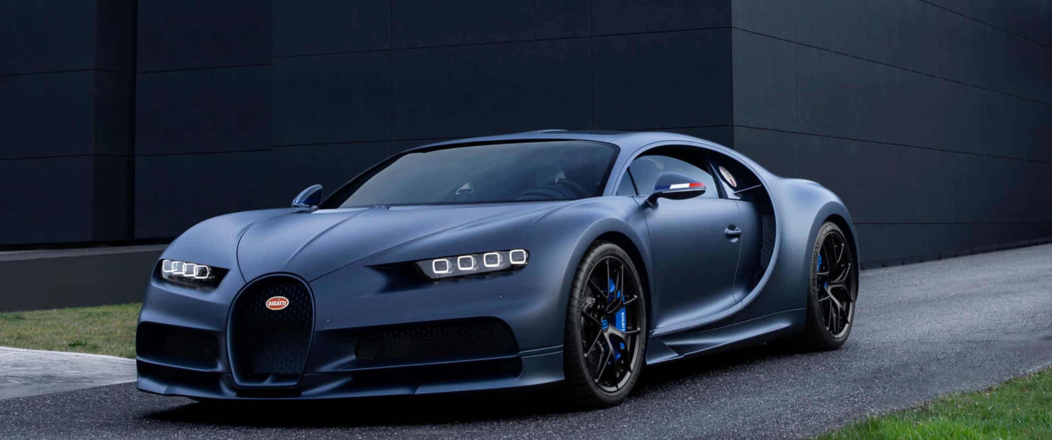 賽車場上的布加迪側影 壁紙 — 一架啞藍色Bugatti Chiron跑車停泊在瀝青路上，旁邊係有草叢嘅深色牆壁。