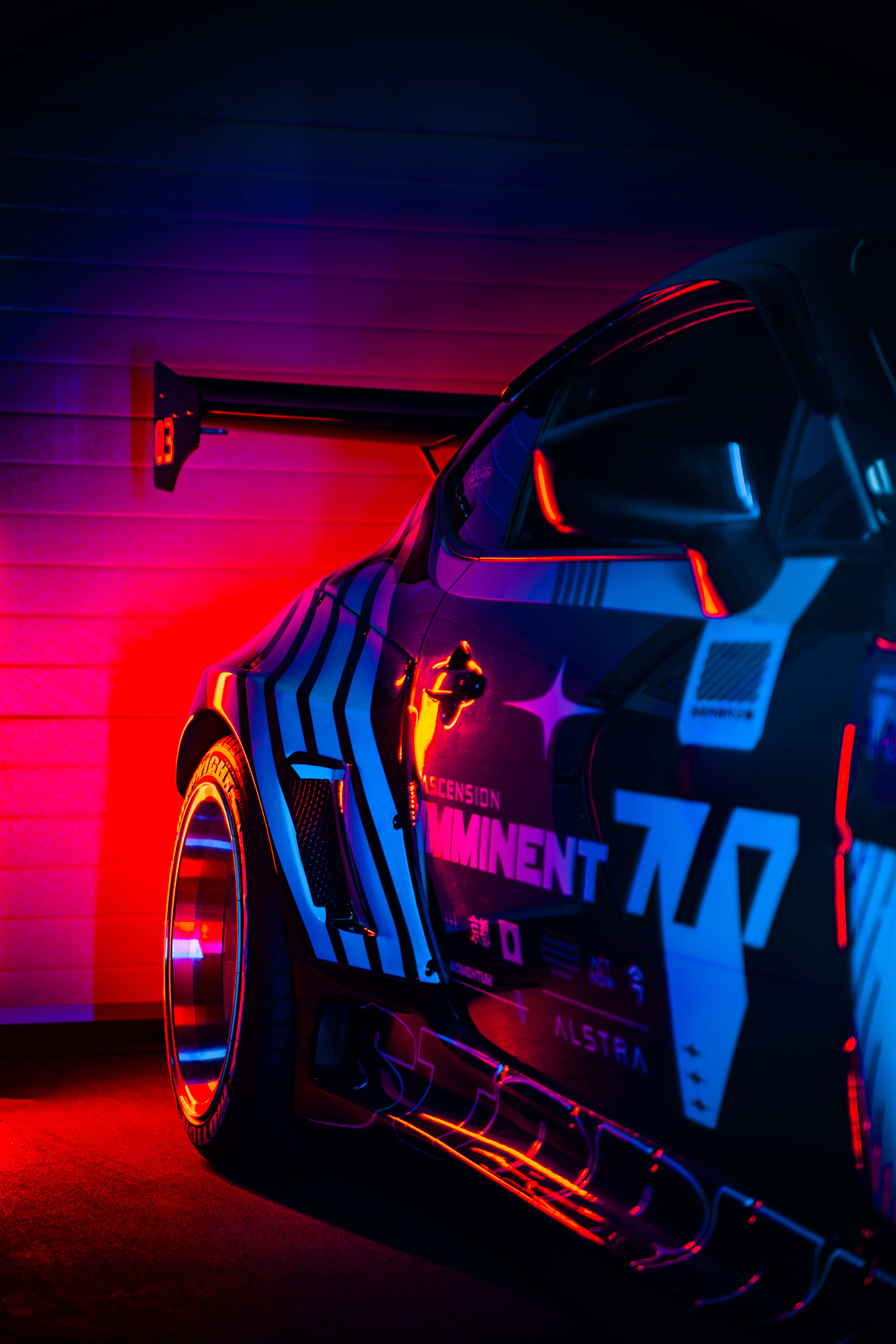 賽車對牆 Pixel 3《Need for Speed Heat》背景 壁紙 — 一輛時尚的黑色賽車，帶有發光的霓虹藍和紅色裝飾，停在昏暗的車庫中，在充滿活力的彩色燈光下。