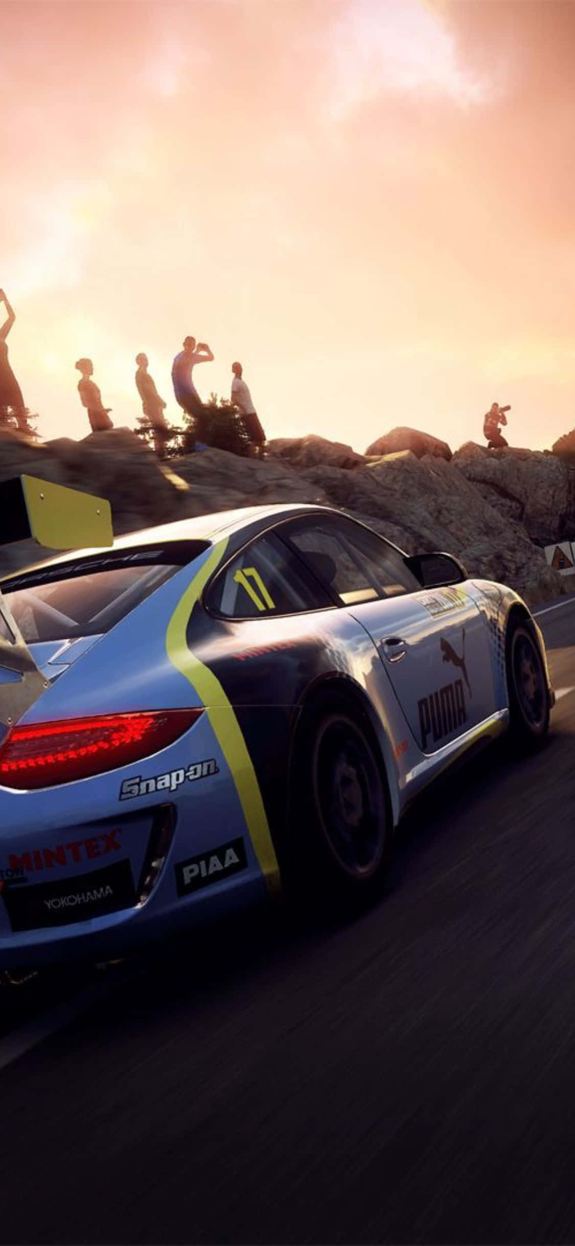 賽車手在《Dirt Rally》比賽中征服彎道 壁紙 — 一輛藍黃色賽車沿著蜿蜒的山路飛馳，觀眾在日落時分從岩石峭壁上觀看。