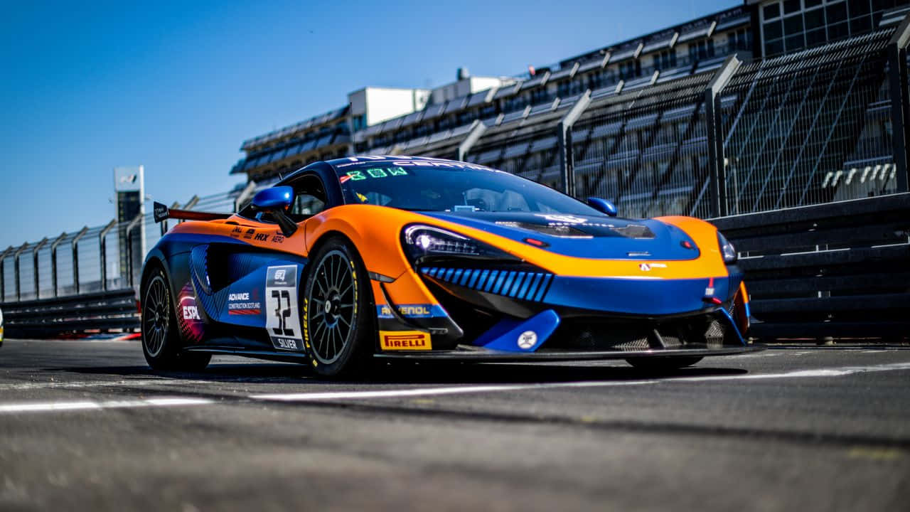 賽車風景 720p 體育背景 壁紙 — 一輛充滿活力的橙色和藍色McLaren賽車在陽光明媚的賽道上疾馳，背景是看台。