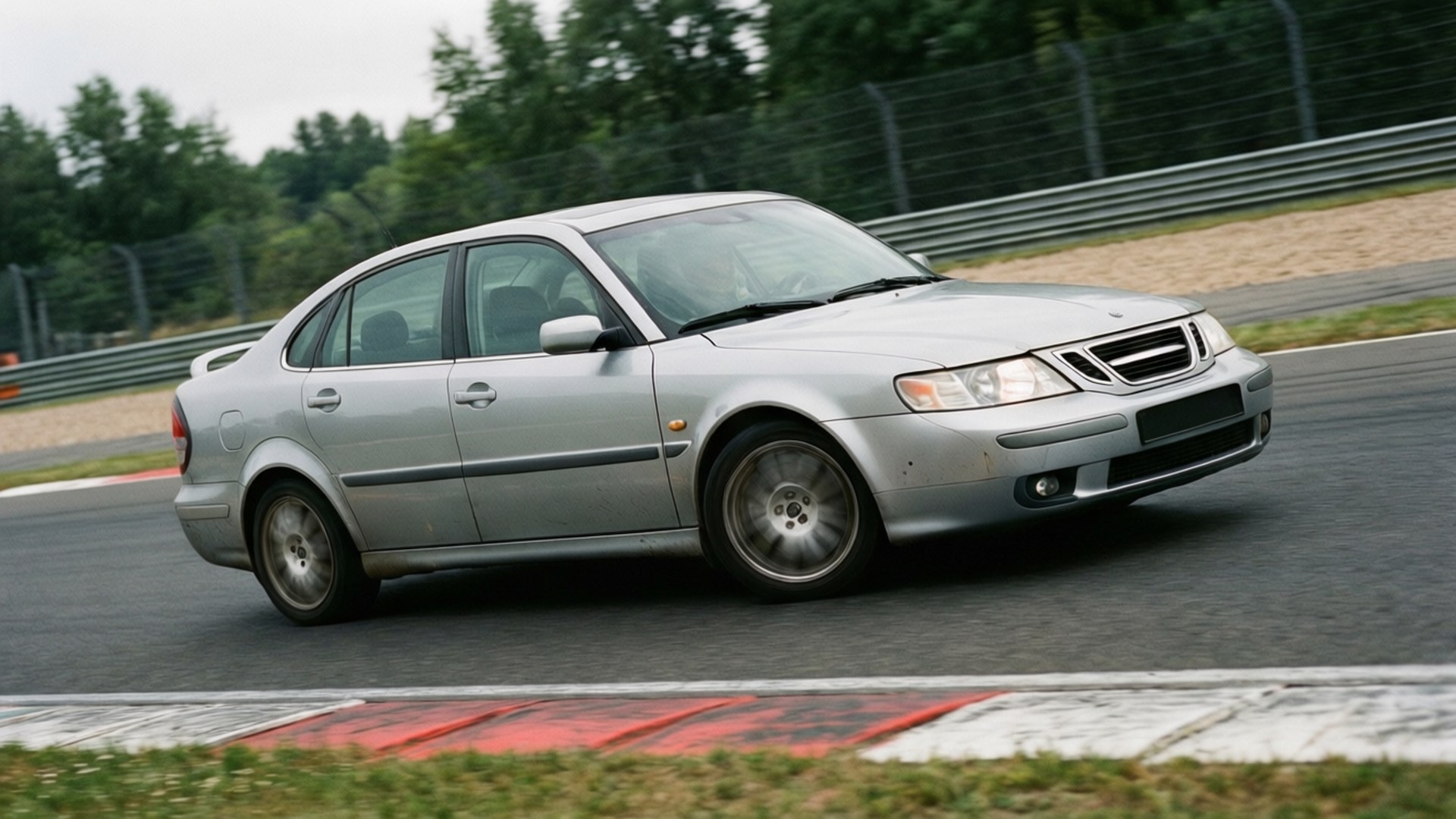 銀色Saab 9-3房車在賽道上疾馳，背景有紅色路緣和綠色樹木。