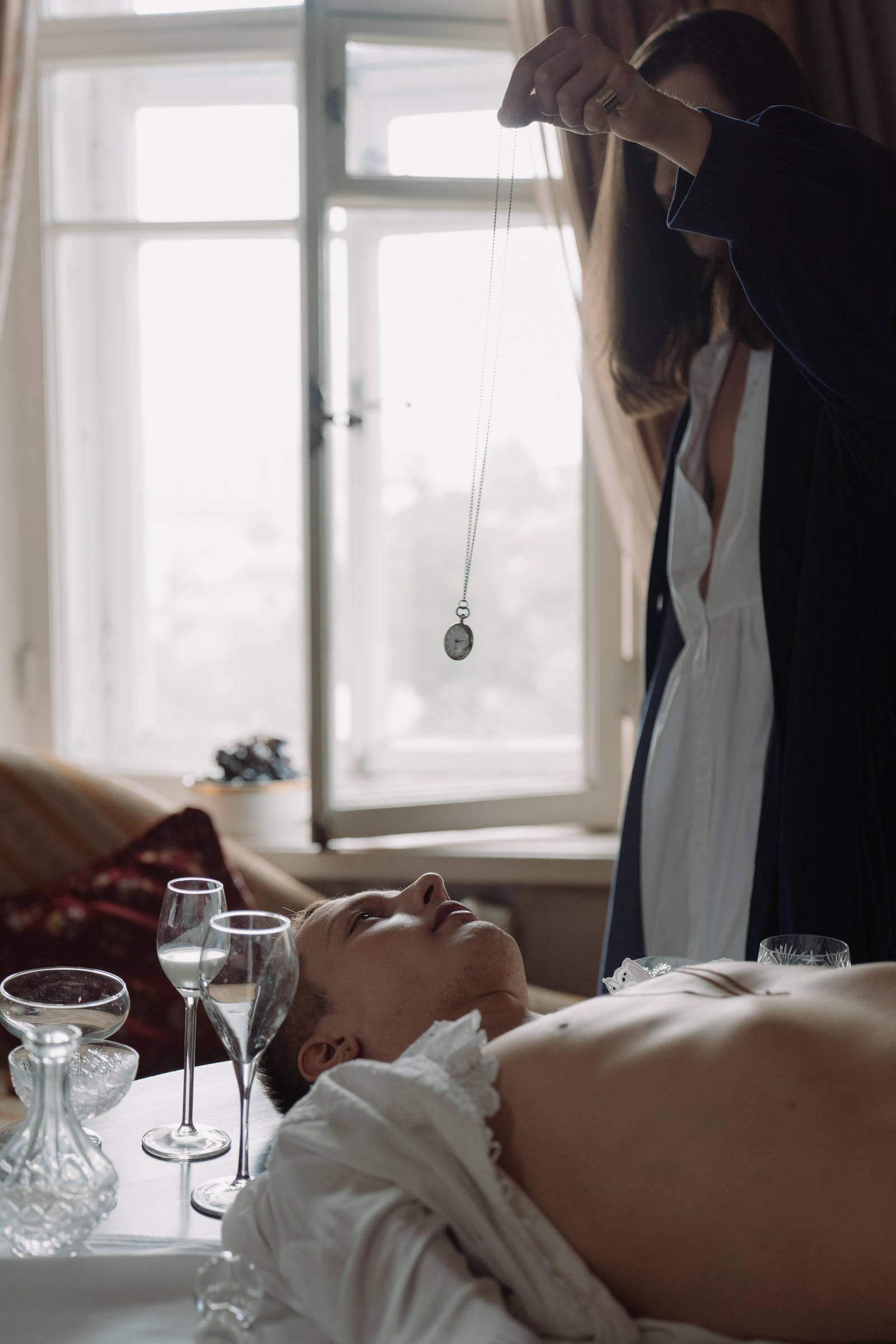 赤裸上身男子接受催眠 壁紙 — 一位身穿深色長袍的女性在陽光充足的房間裡，手持懷錶放在一位斜倚的男子上方，房間裡有酒杯和窗景。