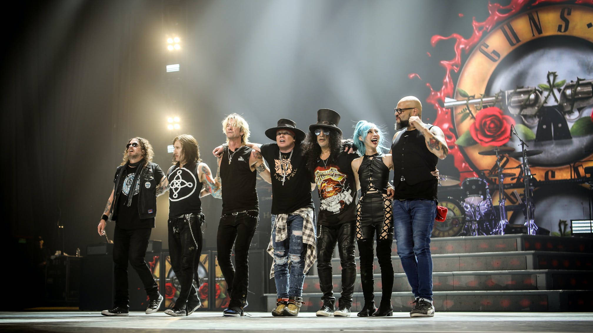 赫希公園音樂節 槍與玫瑰樂隊現場演出 壁紙 — Guns N' Roses 樂隊成員在燈光璀璨的音樂會舞台上自信地擺姿勢，背景是他們標誌性的標誌。