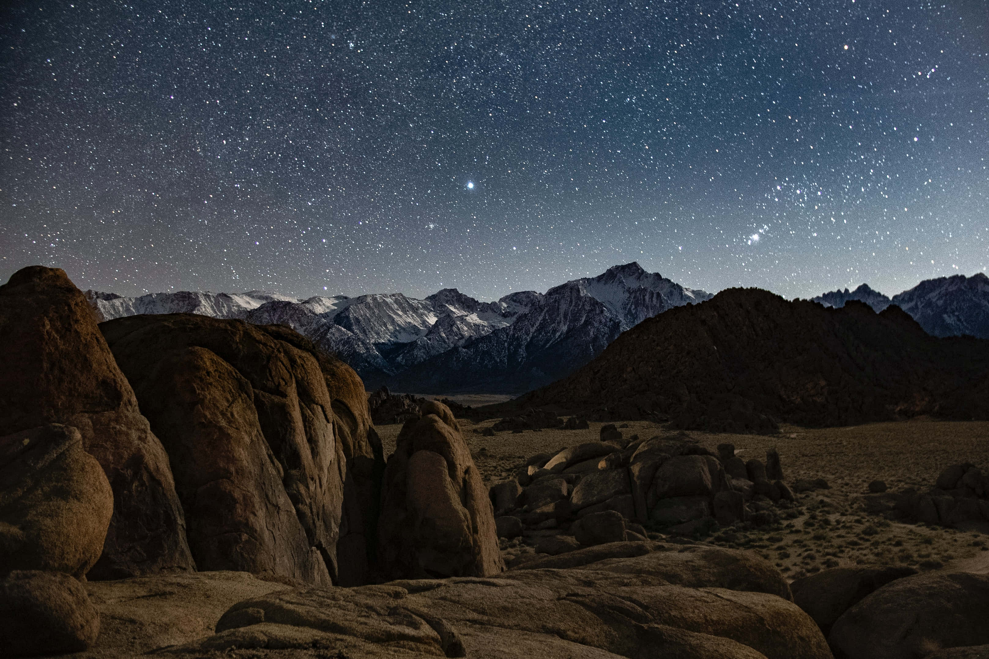 起伏的綠色山丘風景 壁紙 — 深靛藍色的天空下，繁星點點的夜空覆蓋著積雪的山脈和崎嶇的沙漠岩石。