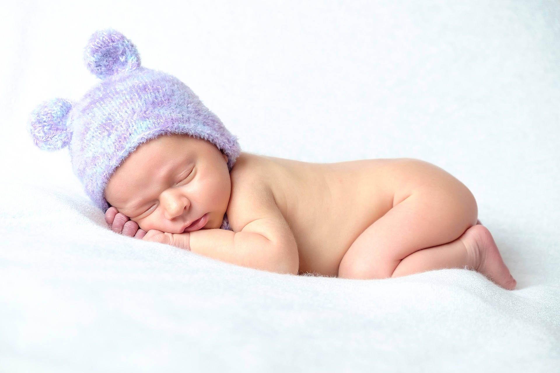 超可愛戴紫帽嬰兒 壁紙 — 一個平靜的新生嬰兒在柔軟的白色毯子上安睡，戴著一頂帶絨球的柔軟薰衣草色針織帽。