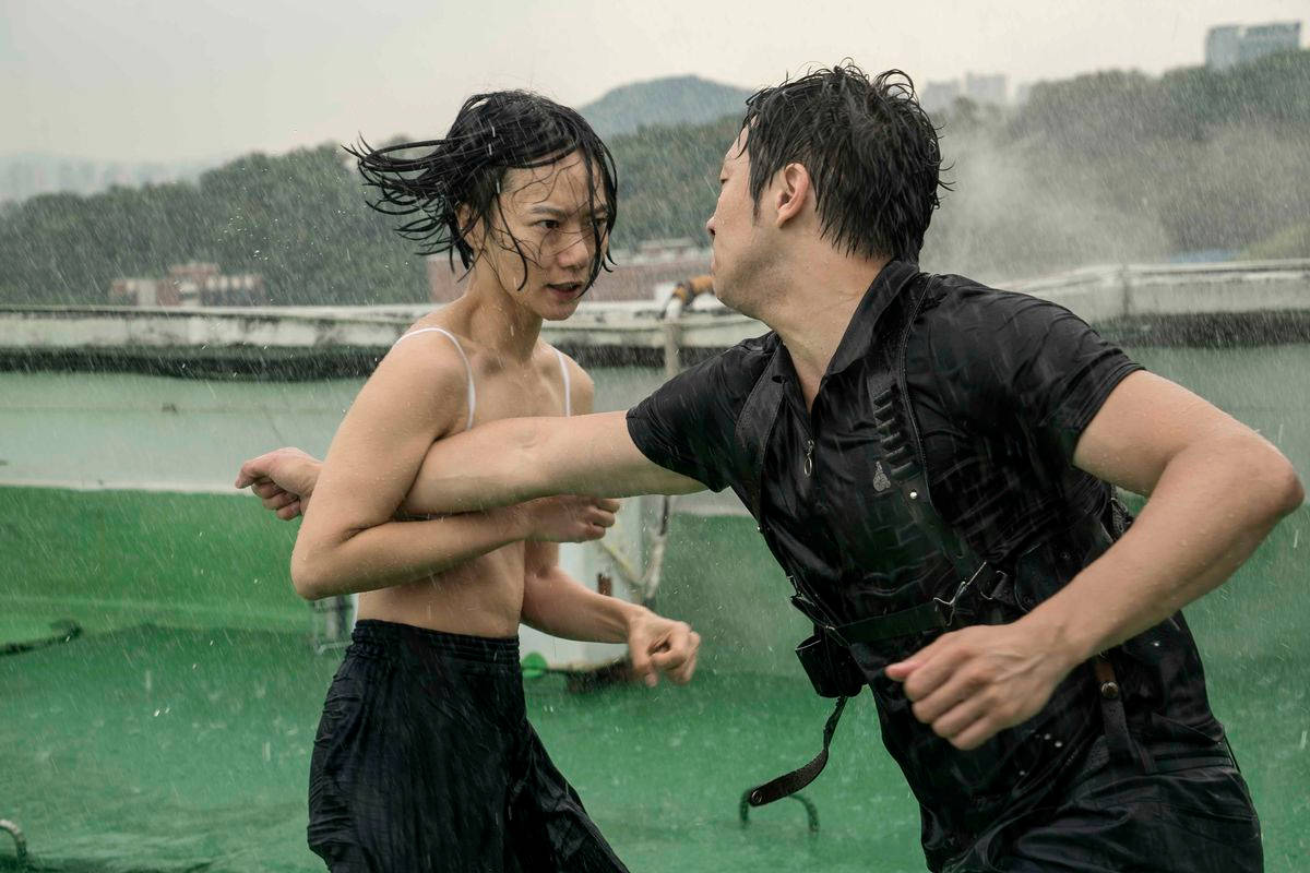 兩個人在雨中在屋頂上打架，身後是城市建築。