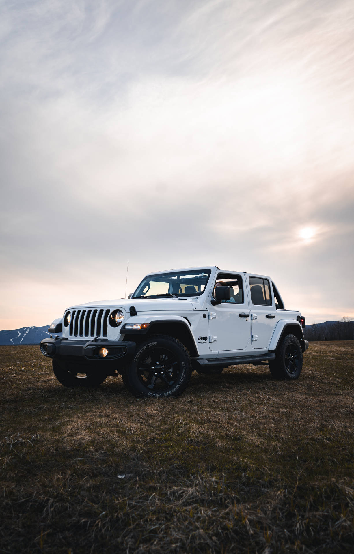 白色Jeep Wrangler停在日落時分的田野上，背景有山脈。
