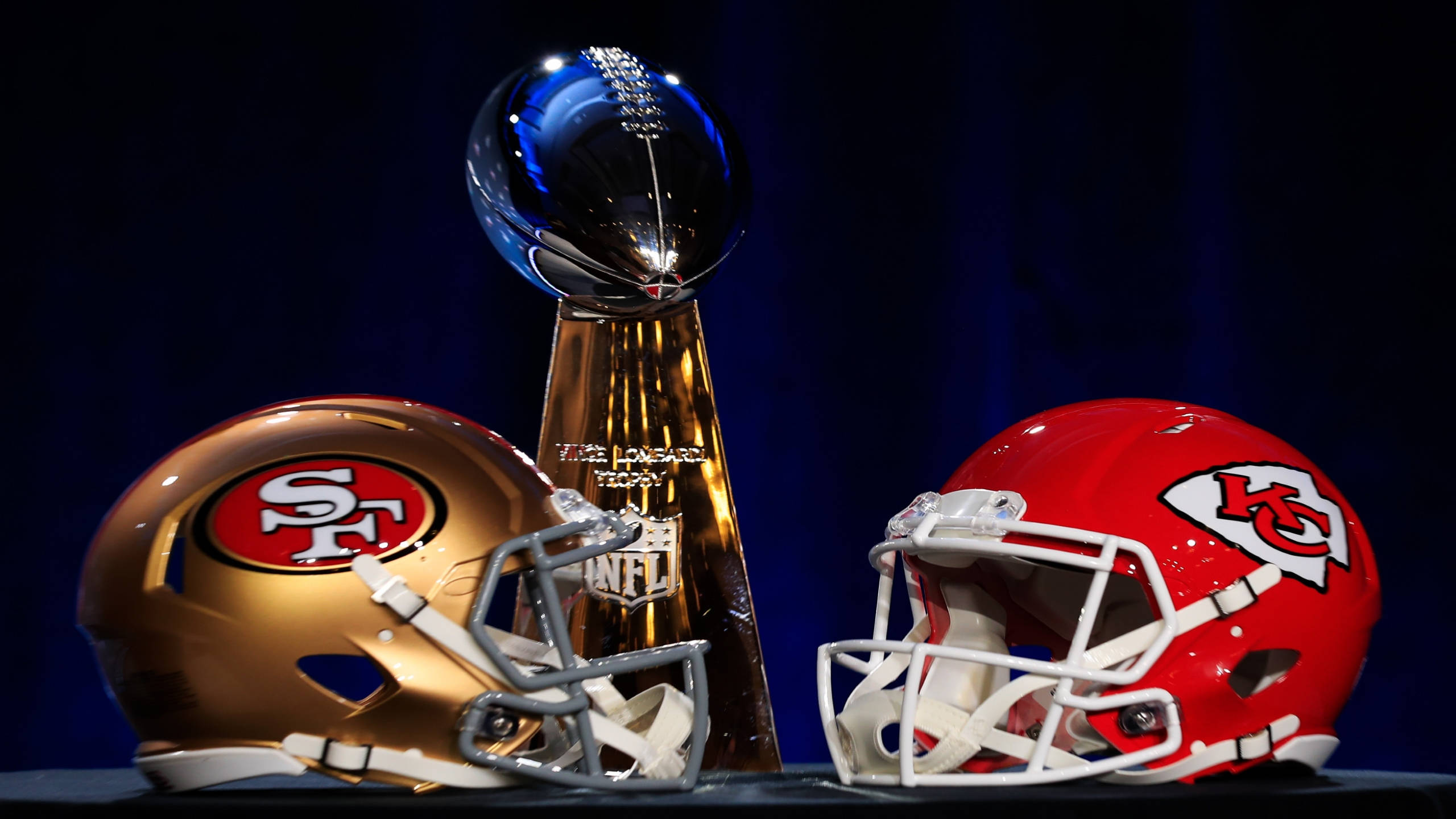 超級碗 2020 羅馬獎盃 壁紙 — 金色和紅色的NFL頭盔在深藍色背景下，簇擁著閃閃發光的隆巴迪獎盃。