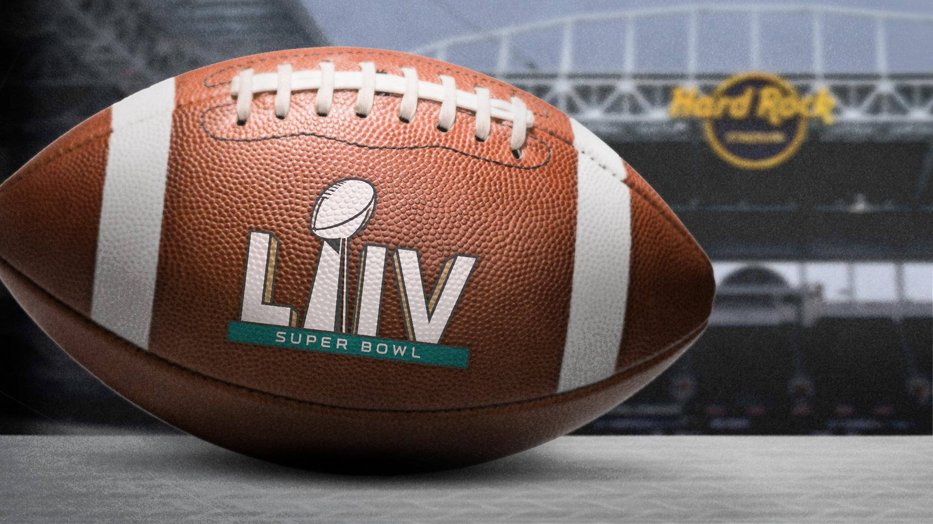 超級碗LIV足球 壁紙 — 近距離拍攝一個棕色皮革 Super Bowl LIV 足球，帶有白色鞋帶和標誌，放置在灰色表面上，背景是模糊的體育場。