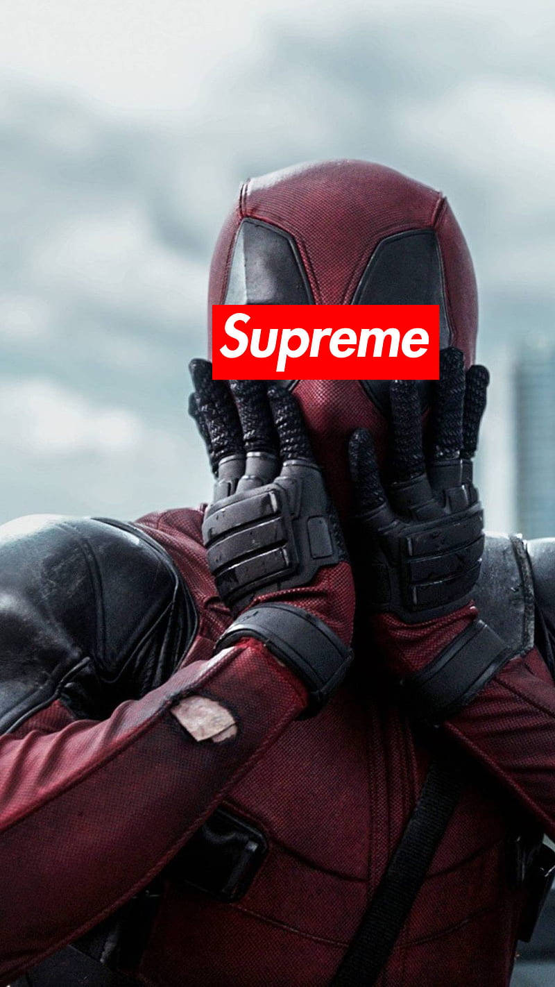 超級英雄 Supreme 死侍驚訝表情 壁紙 — 死侍穿著紅黑戰衣，雙手遮臉，露出紅色Supreme標誌，背景是多雲的天空。