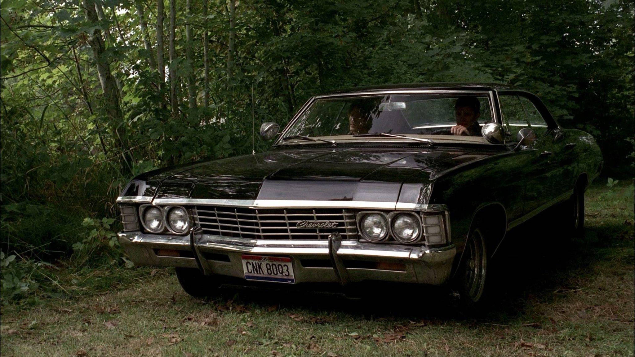 超自然1967款雪佛蘭Impala 壁紙 — 一輛黑色經典雪佛蘭敞篷車停放在茂密的綠色森林中，車內有兩個人。