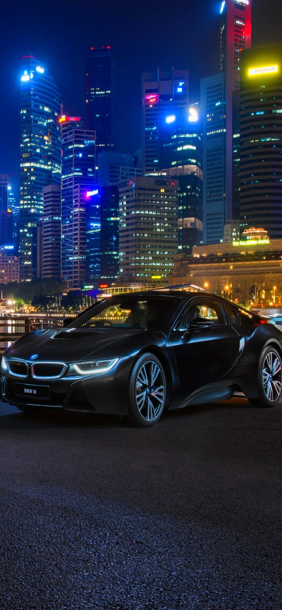 超越完美的魅力：iPhone X與BMW 壁紙 — 一輛時尚的黑色BMW i8跑車在夜晚的霓虹燈下，以高聳的摩天大樓為背景閃閃發光。