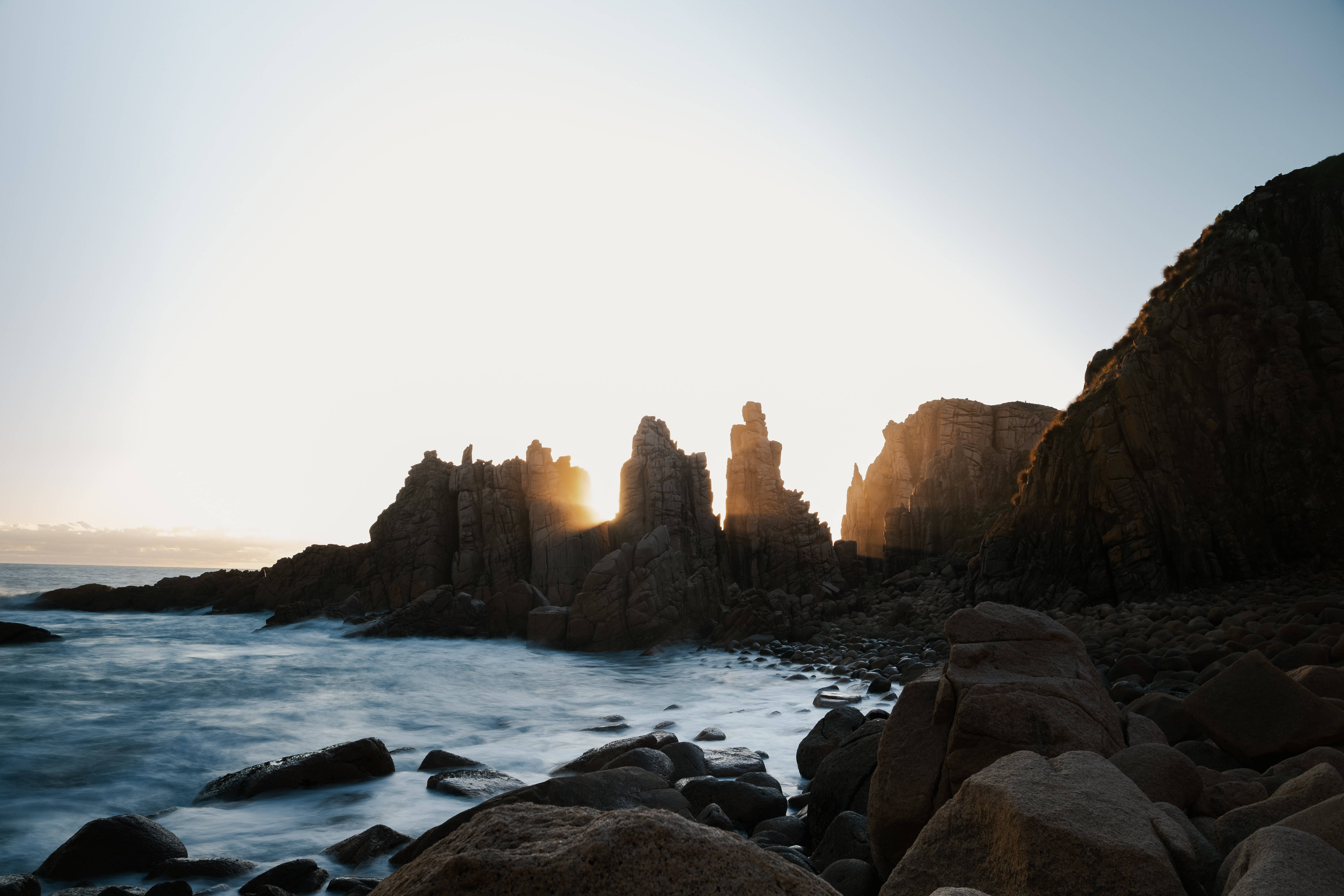 超酷的尖峰景觀 壁紙 — 夕陽的光線穿過參差不齊的海崖，海浪輕輕拍打著暗黑的岩石，呈現寧靜的海岸景象。