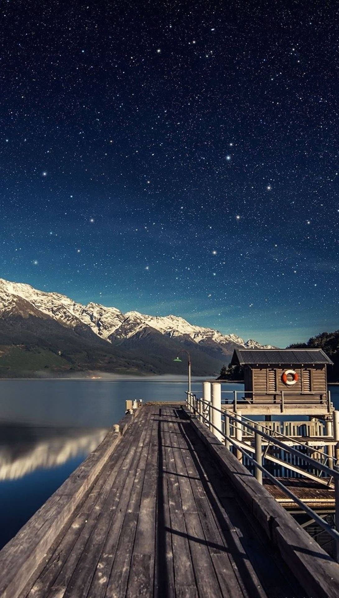 夜晚的木製碼頭，星星和雪山倒映在平靜的水面上。