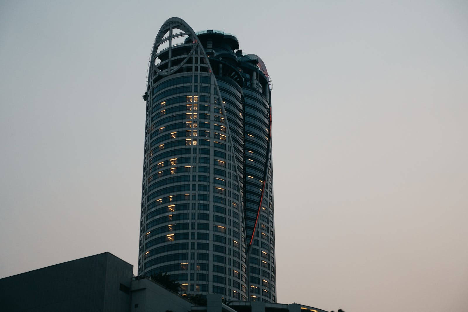超高解析度摩天大樓 壁紙 — 一座高聳的現代摩天大樓，擁有弧形玻璃幕牆和亮著的窗戶，矗立在昏暗的天空下。