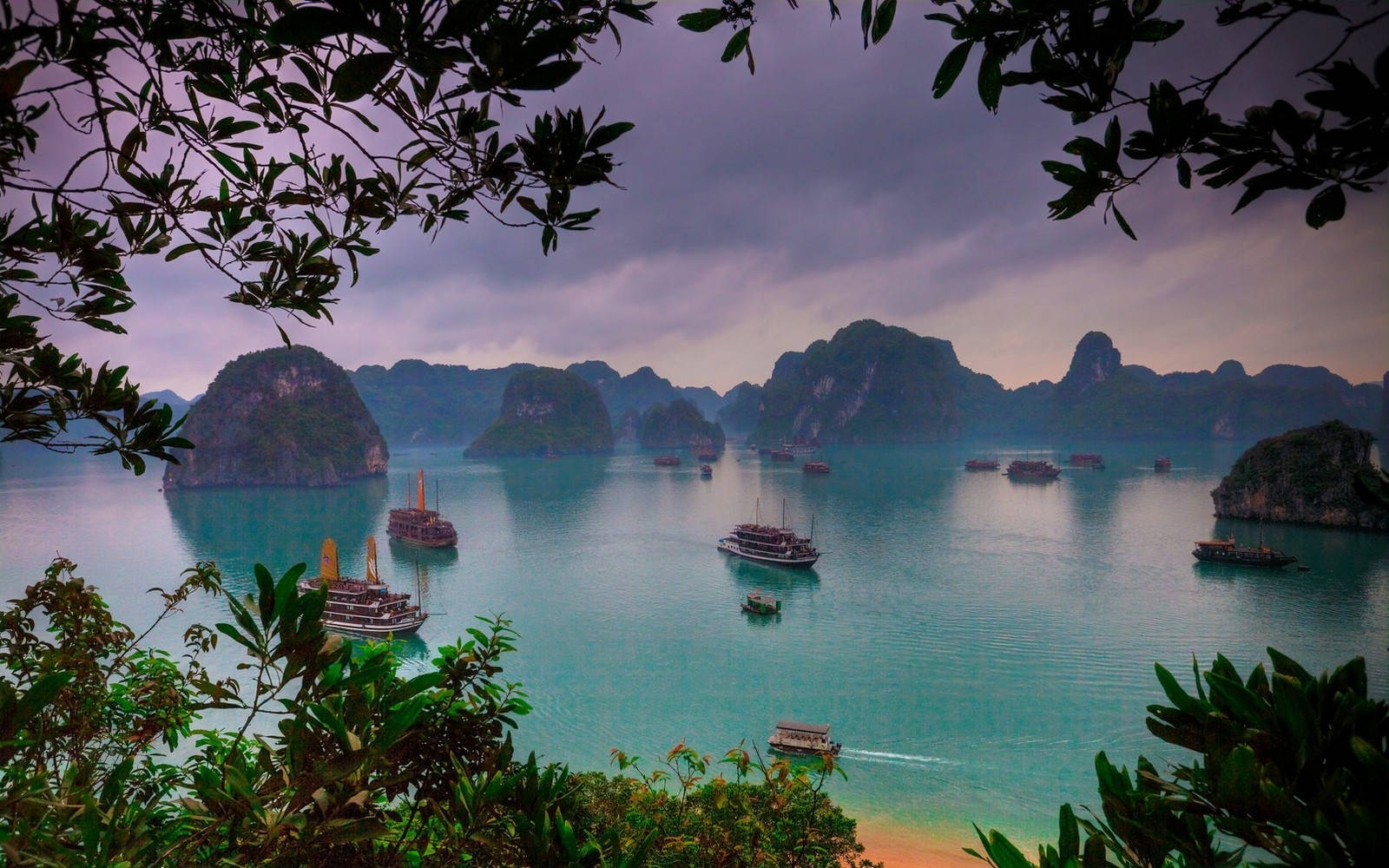 越南下龍灣風景 壁紙 — 一個寧靜的熱帶海灣，被茂密的樹葉環繞，石灰岩喀斯特地貌和傳統船隻在陰沉天空下漂浮在碧綠的海水上。