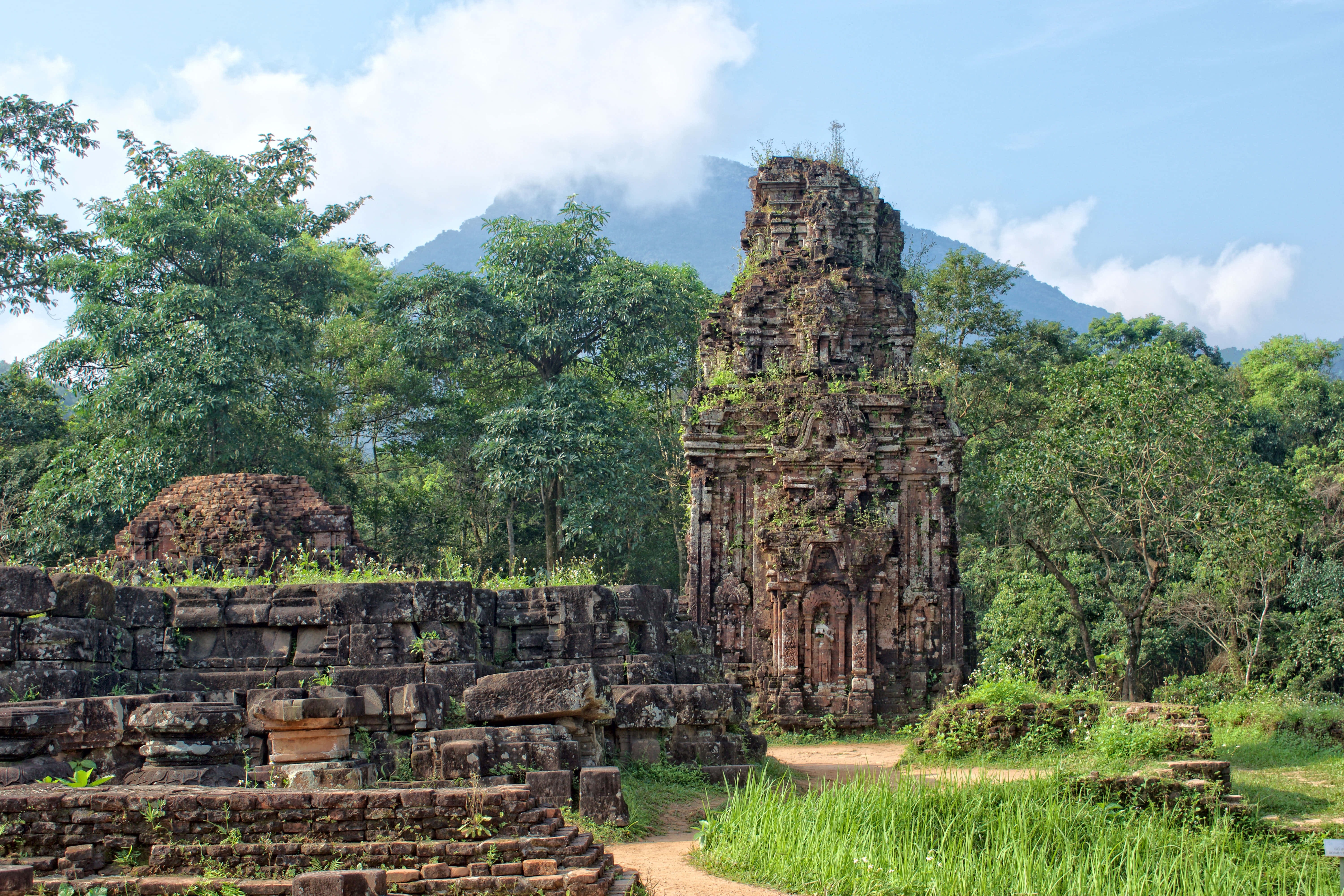 越南美山聖地的古建築 壁紙 — 被綠色植物覆蓋的古老石廟遺跡，在明亮的藍天下矗立在霧氣繚繞的山前。