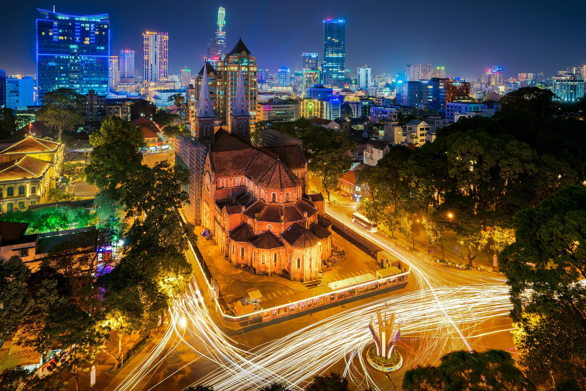 越南聖母院 壁紙 — 充滿活力的夜景，一座歷史悠久的教堂在溫暖燈光下閃耀，周圍是發光的摩天大樓和車流燈跡。