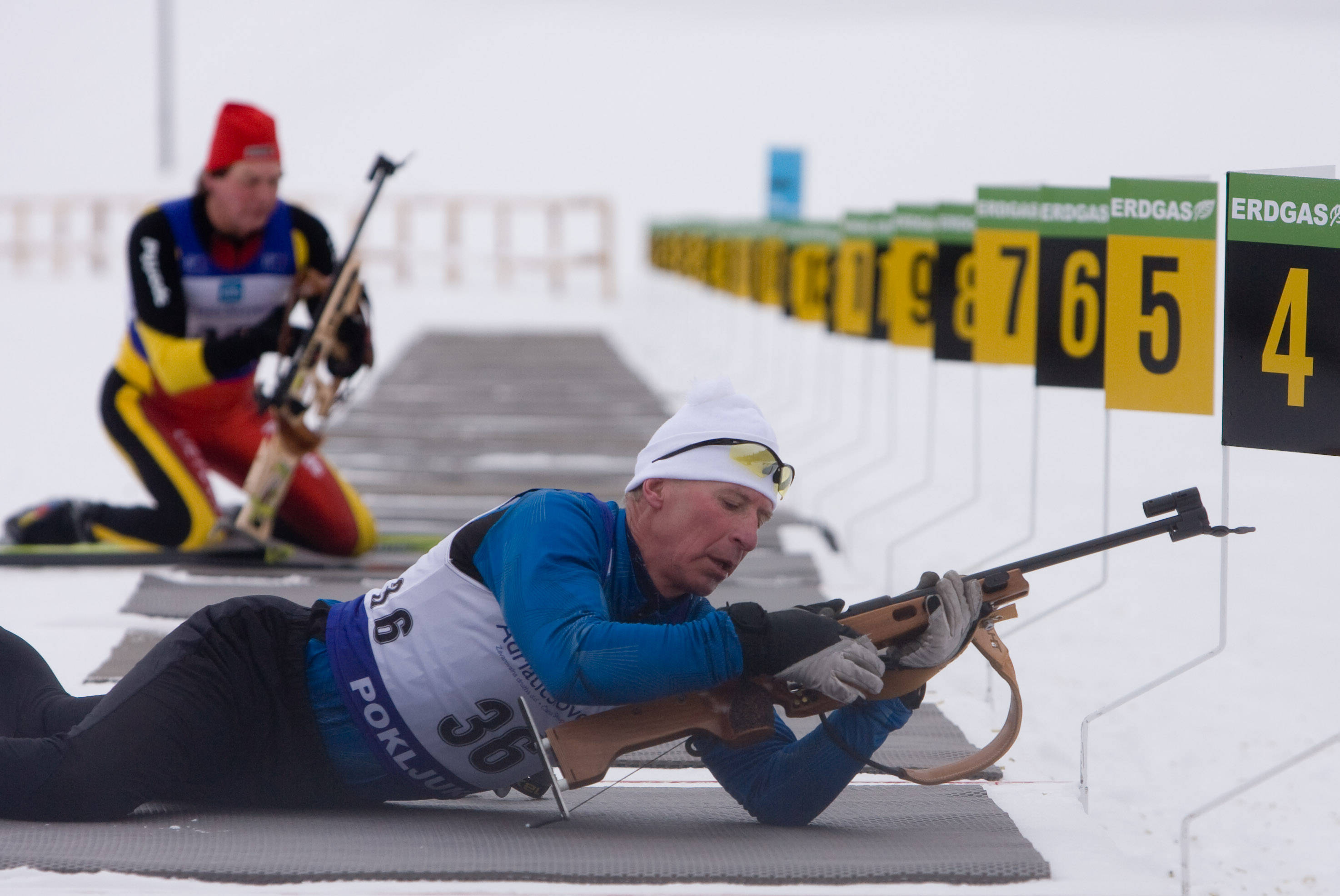 越野滑雪射擊選手臥姿裝填步槍 壁紙 — 冬季裝備的冬季兩項運動員在雪地中以俯臥姿勢向帶有黃色和黑色數字的目標射擊步槍。