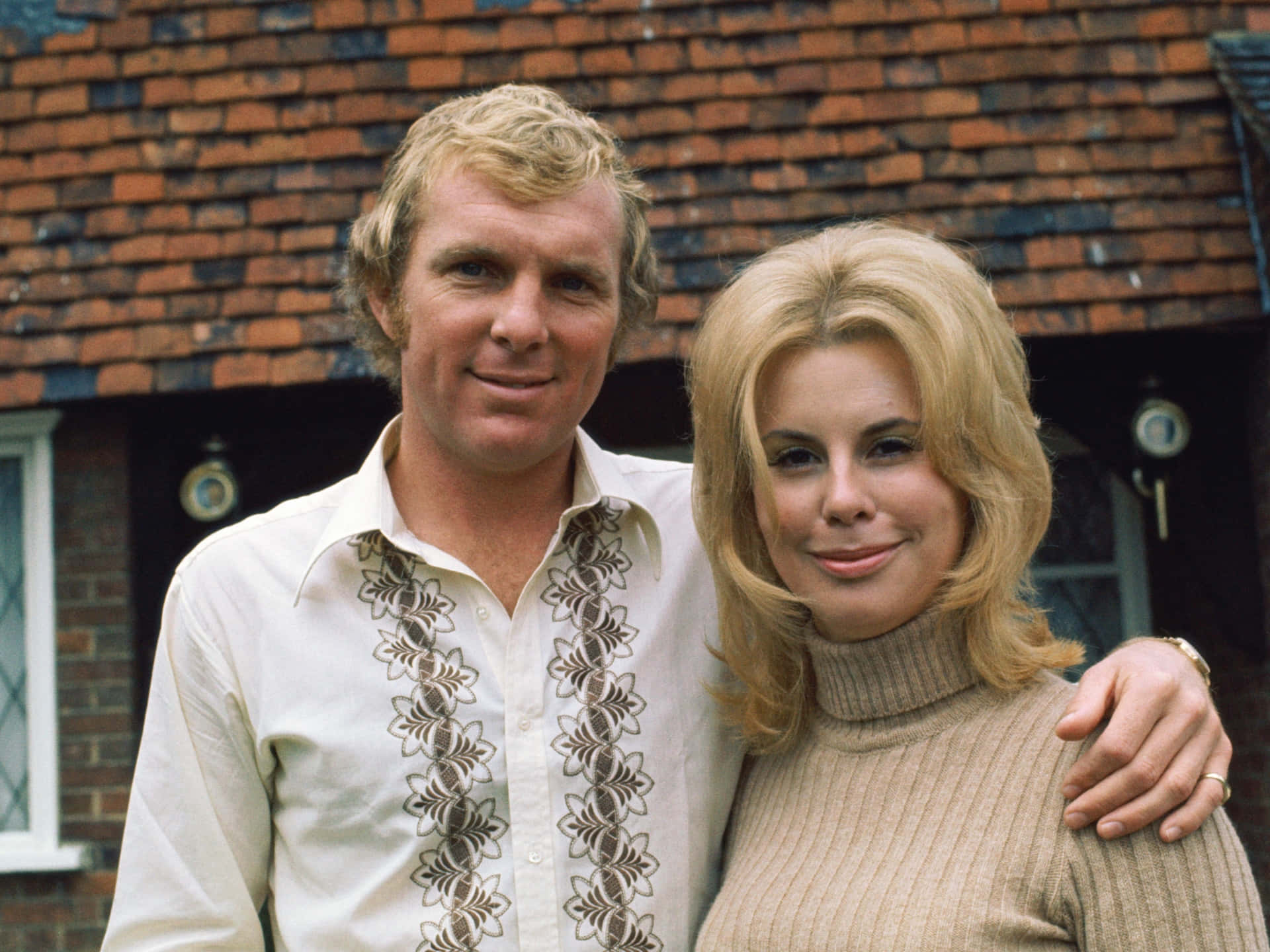 足球員Bobby Moore與Tina Moore的甜蜜合照 壁紙 — 一對微笑的金髮情侶穿著1970年代時尚，在磚砌建築和瓦片屋頂前溫馨地擺姿勢。
