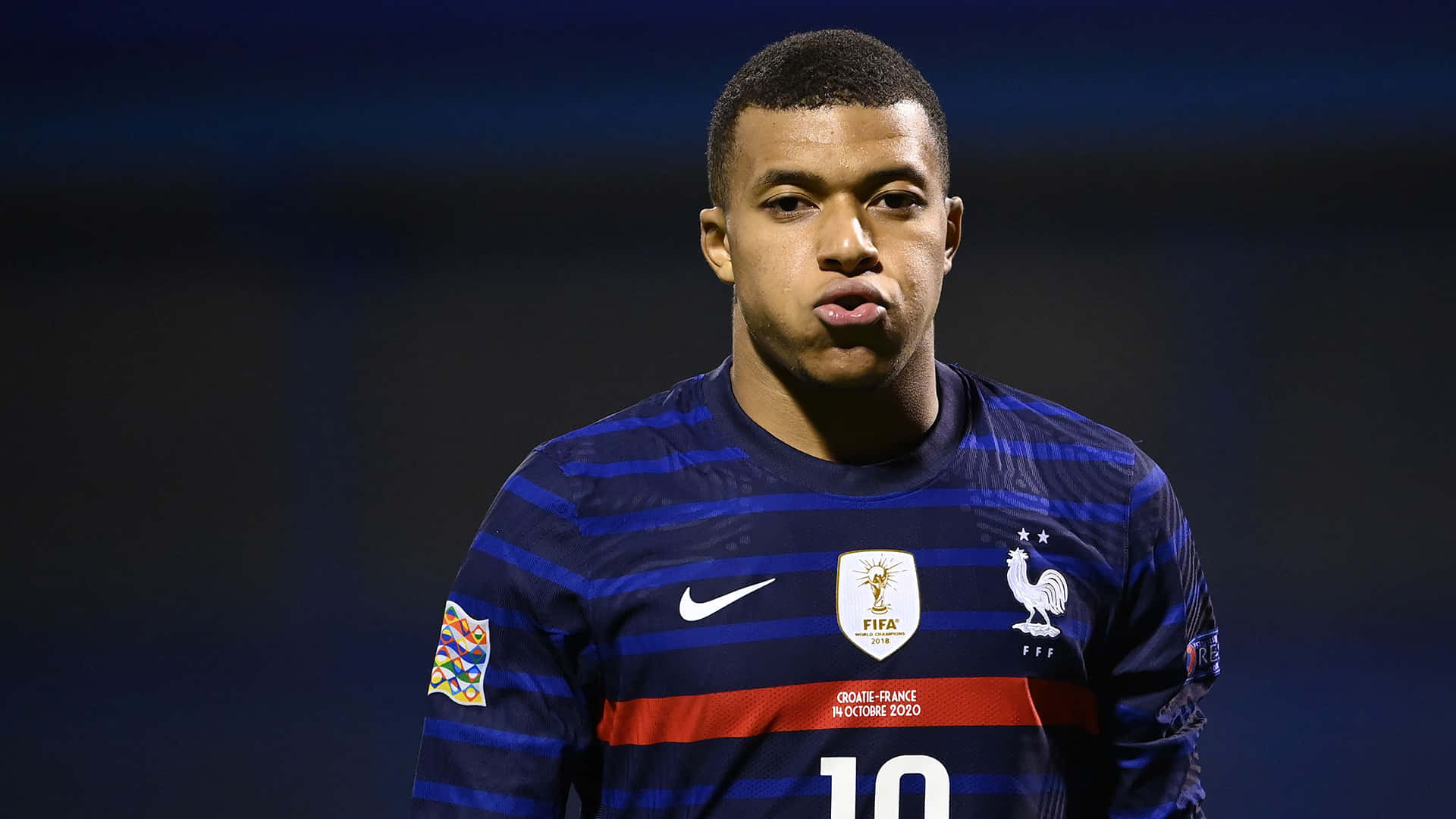 Mbappe 桌布 壁紙 — 足球員基利安·麥巴比在球場燈光下穿著法國藍色間條國家隊球衣，嘟起嘴唇的特寫。