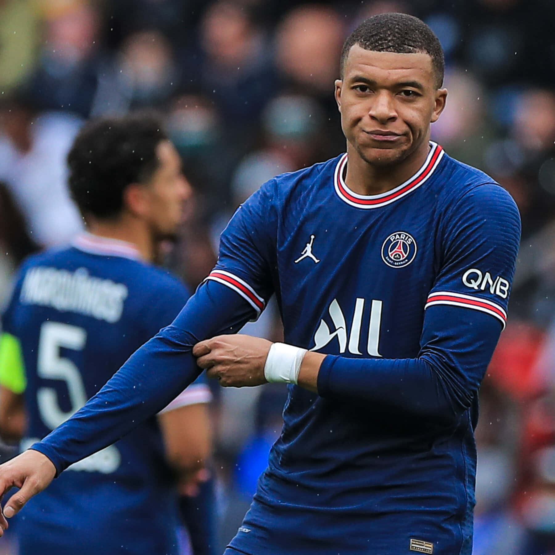 Mbappe 桌布 壁紙 — 足球員基利安·麥巴比身穿藍色PSG球衣，在日間比賽中自信微笑，背景是模糊的球場人群。