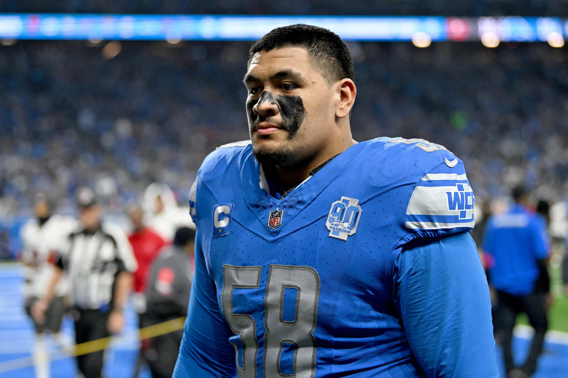 足球員場上風采 壁紙 — 一位專注的NFL線衛，身穿藍色58號球衣，臉上塗有黑色顏料，並帶有隊長'C'臂章，在比賽中站在場上。