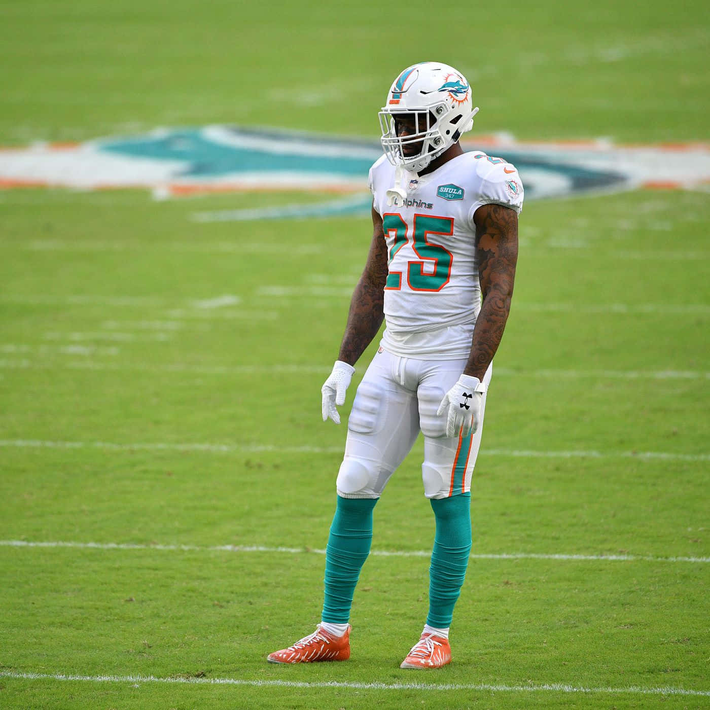 足球場上的Xavien Howard球員 壁紙 — 一名身穿白色和青綠色球衣的邁阿密海豚隊美式足球員在日光下獨自站在充滿活力的綠色球場上。