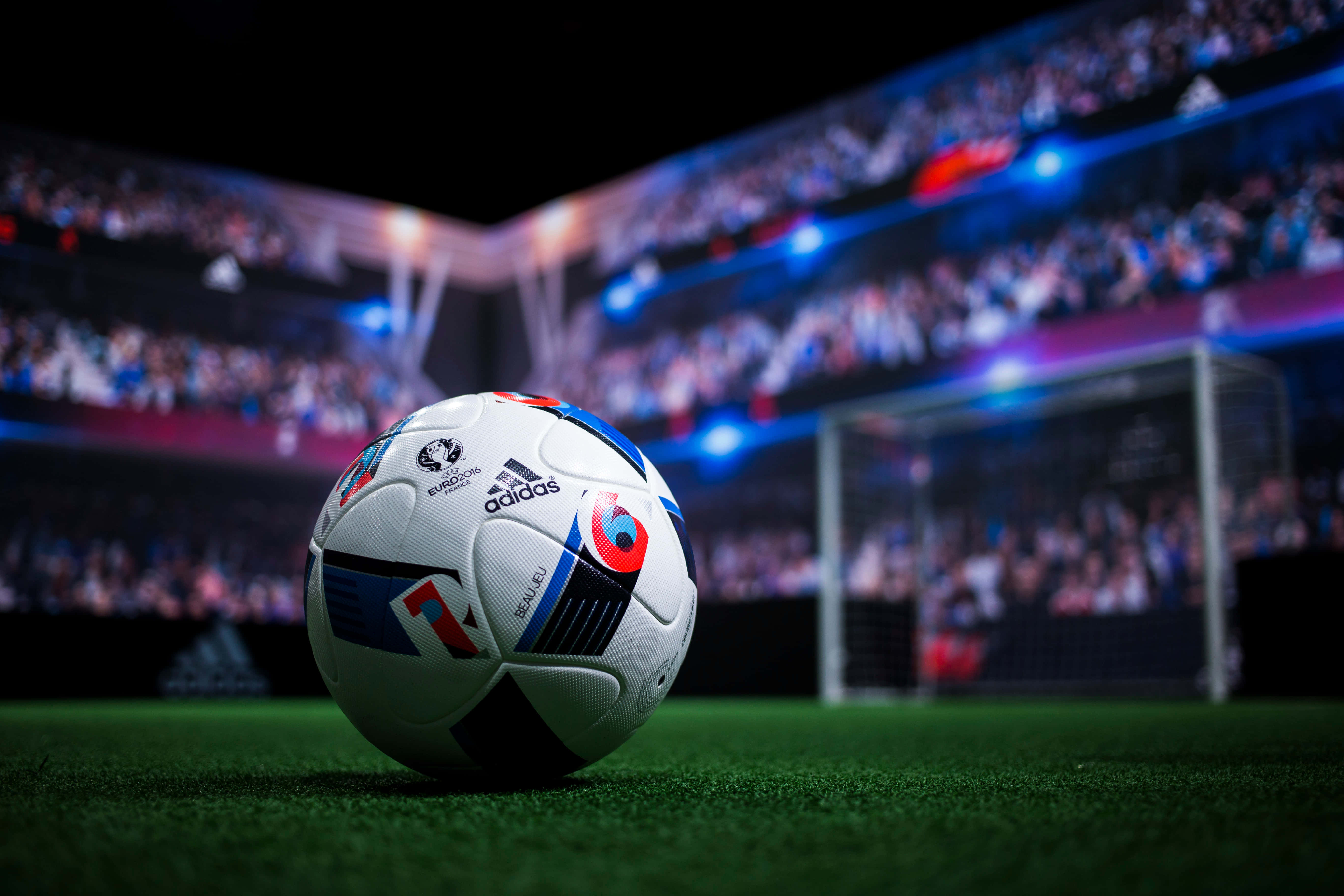 足球場背景：足球與觀眾 壁紙 — 一個UEFA Euro 2020足球放在充滿活力的綠色草地上，模糊的體育場人群和球門網在戲劇性的藍色燈光下閃耀。