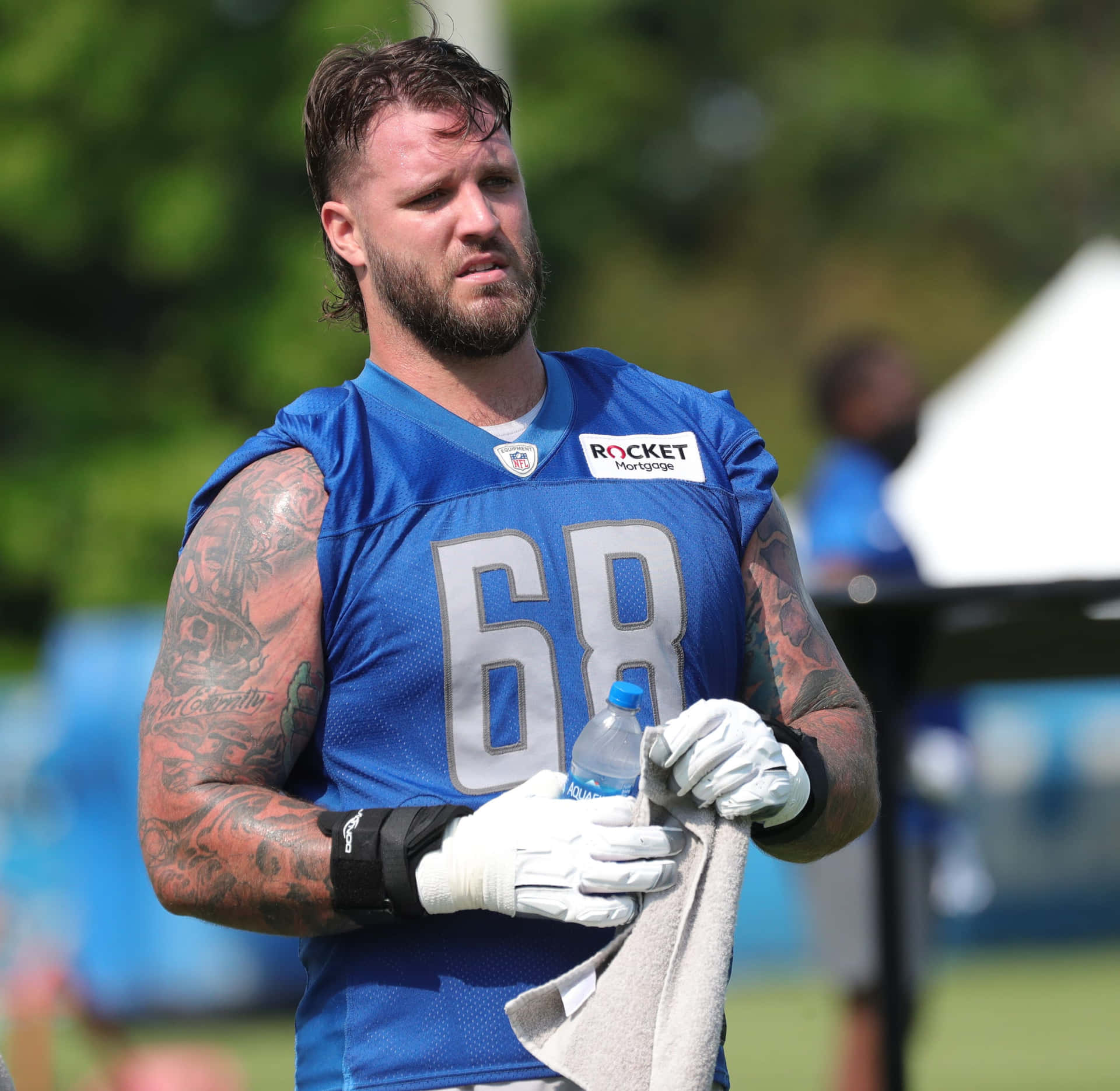 足球攻防鋒線運動員泰勒·德克爾 壁紙 — 一位專注的NFL球員身穿藍色球衣，號碼68，在陽光明媚的戶外訓練中用毛巾擦汗。
