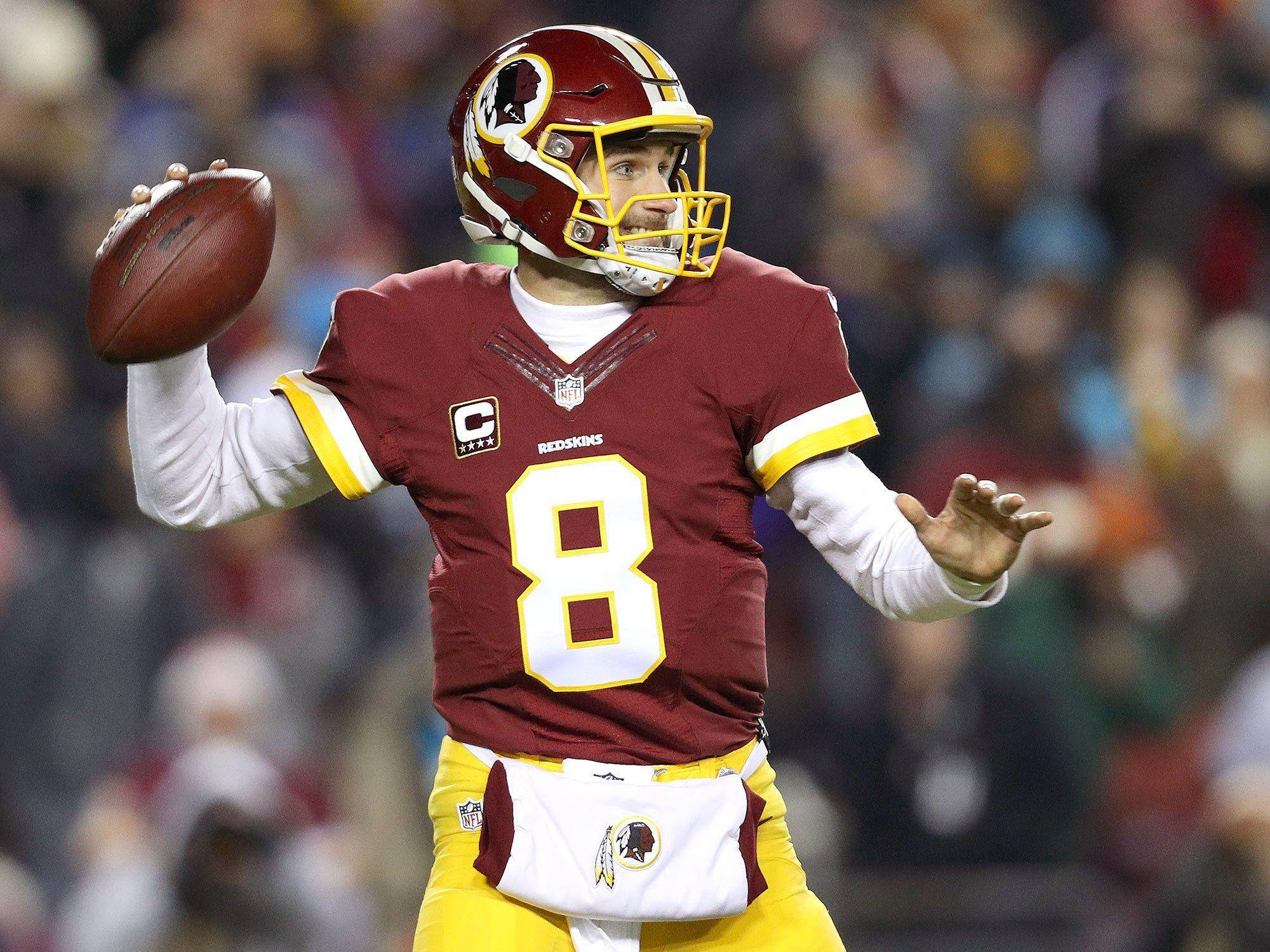 足球明星 Kirk Cousins 壁紙 — 華盛頓紅皮隊的四分衛身穿栗色和金色制服，在體育場燈光下的夜間比賽中準備投擲橄欖球。