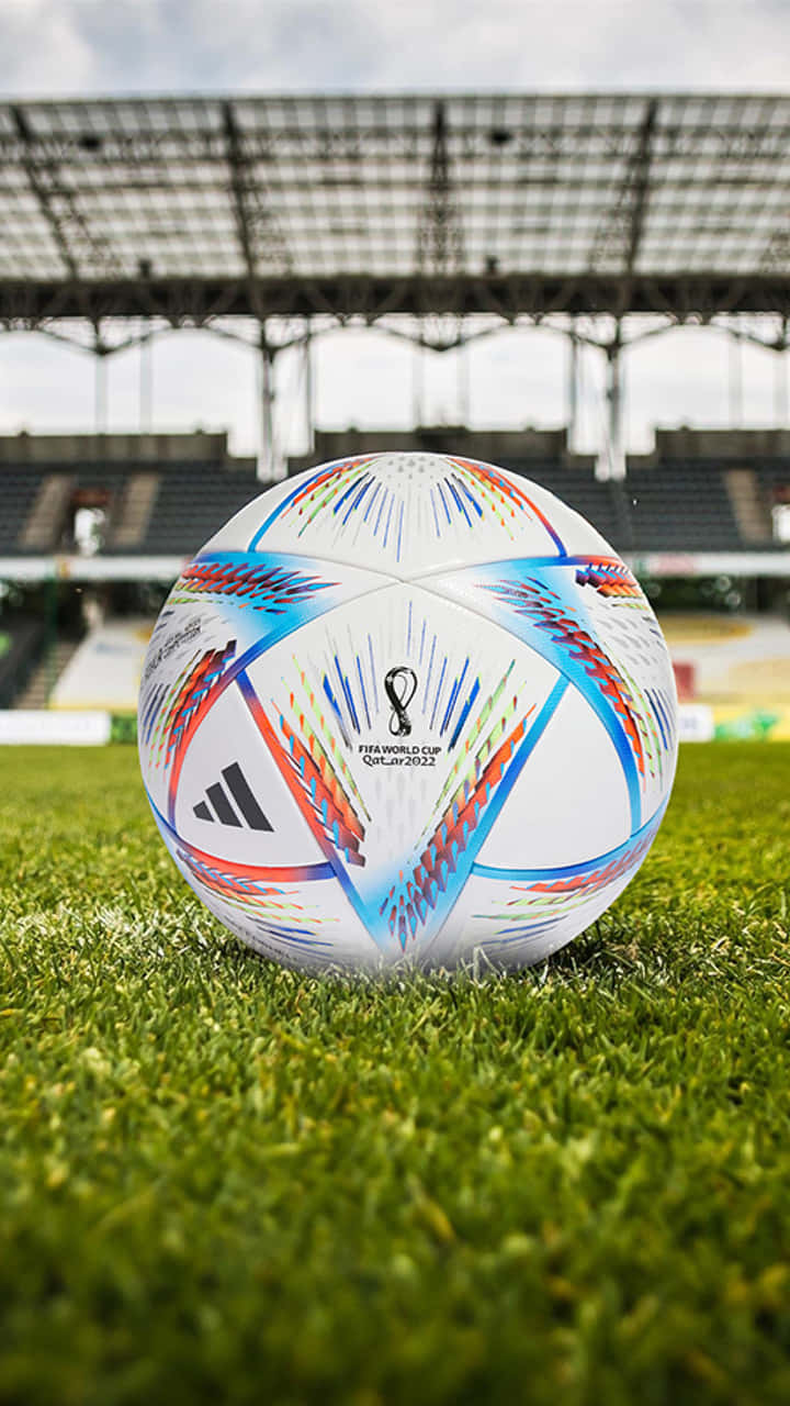 足球背景：繽紛色彩 壁紙 — 一個充滿活力的FIFA世界盃卡塔爾2022足球，放在陰天下一片空蕩蕩的體育館內茂密的綠草上。