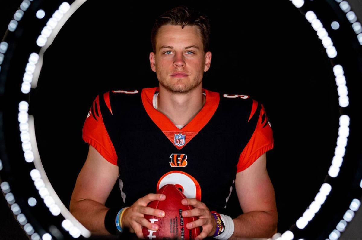 足球運動員 Joe Burrow 壁紙 — 一名身穿孟加拉虎隊球衣的美式足球四分衛，在黑色背景上，手持一個被發光圓形光環框住的球。