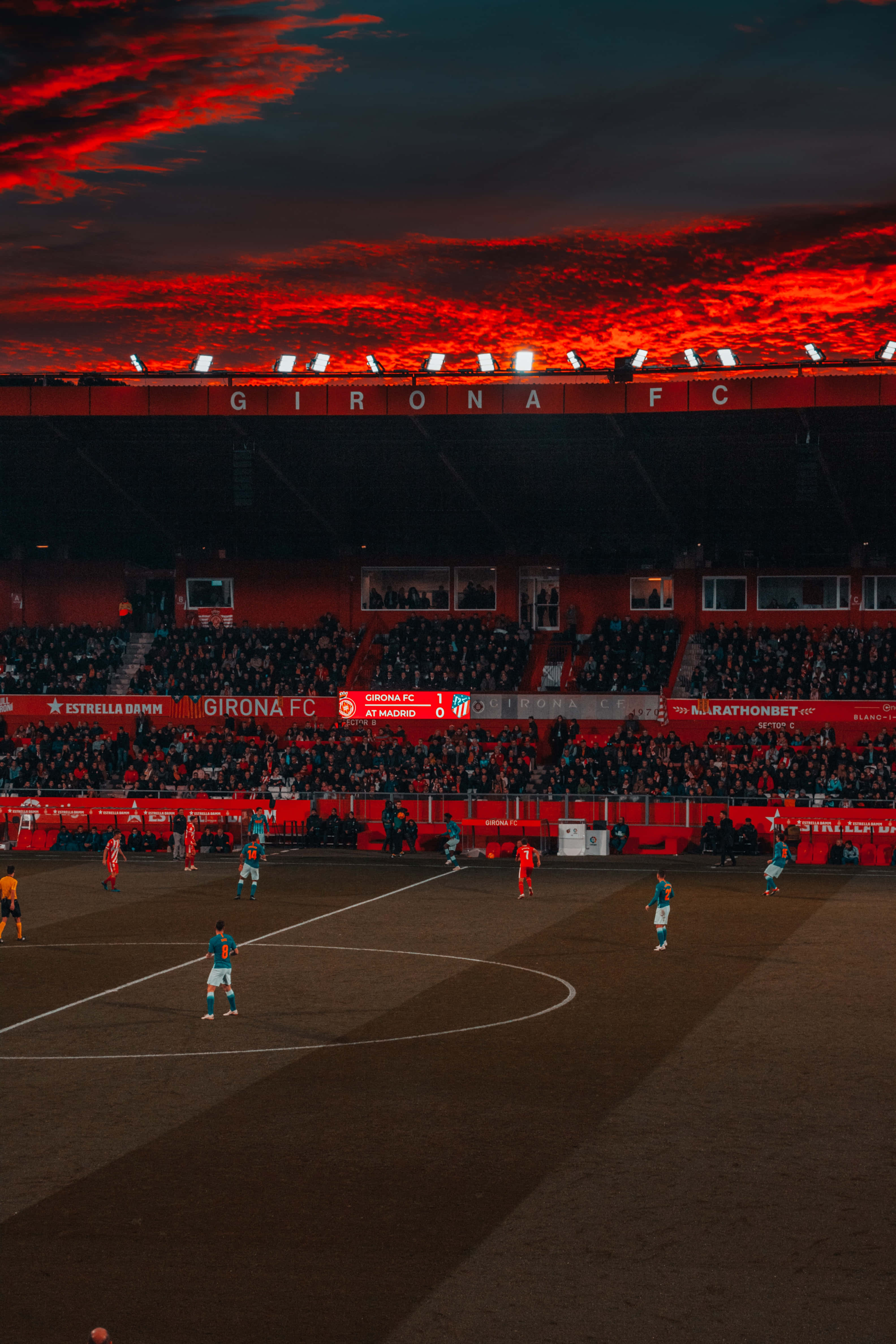 Soccer Stadium 桌布 壁紙 — 足球運動員在吉羅納FC球場，於戲劇性的紅色夕陽天空下，在擠滿球迷的球場中競技。