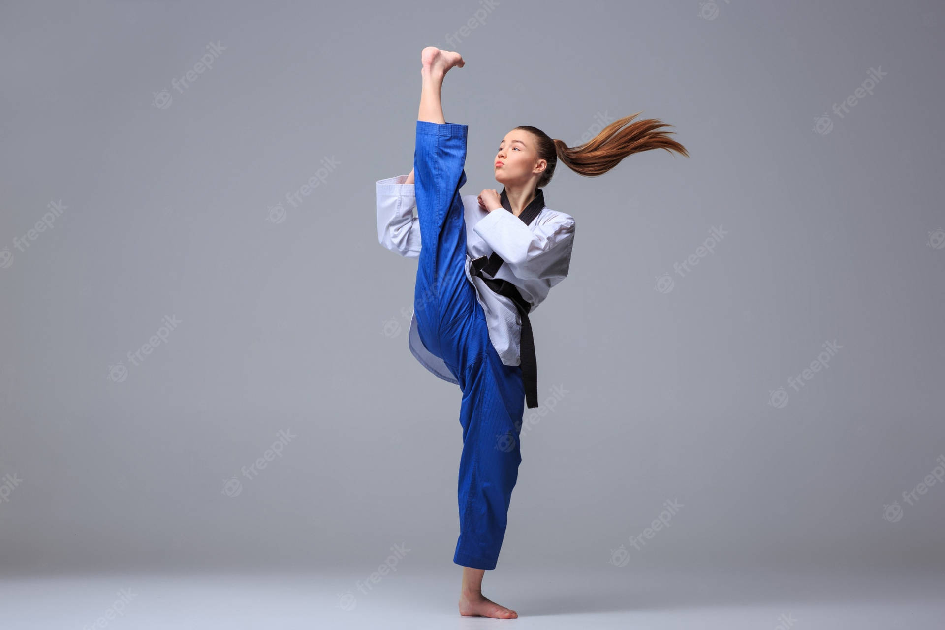 跆拳道女性黑帶運動員 壁紙 — 一位身穿藍色道服的女武術家，在純灰色工作室背景前，秀髮飄逸，做出高踢動作。