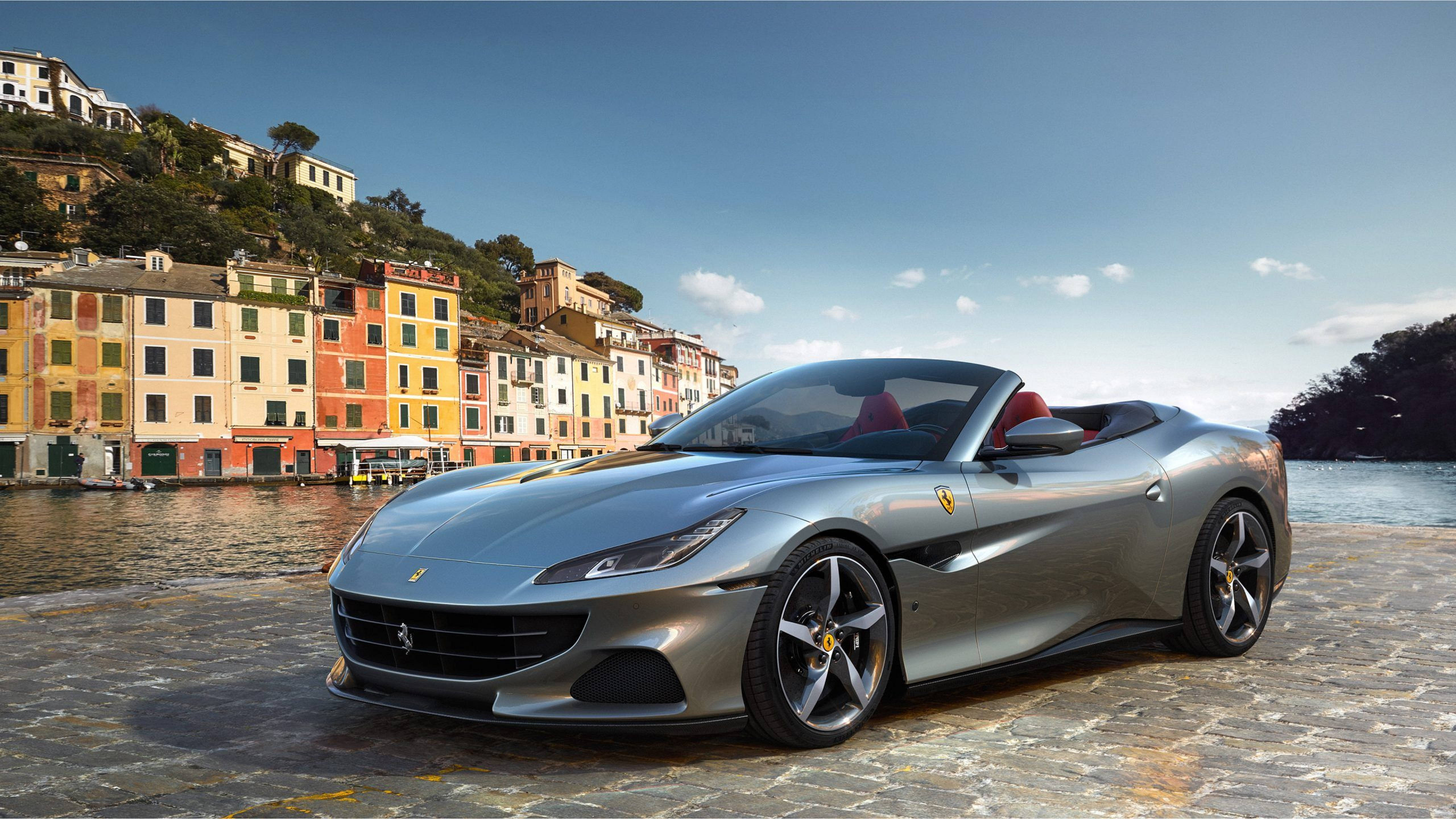 跑車法拉利Portofino 壁紙 — 一架流線型銀色法拉利 Portofino 開篷車停泊在鵝卵石海濱，背景是色彩繽紛的意大利山丘建築和寧靜的水面。
