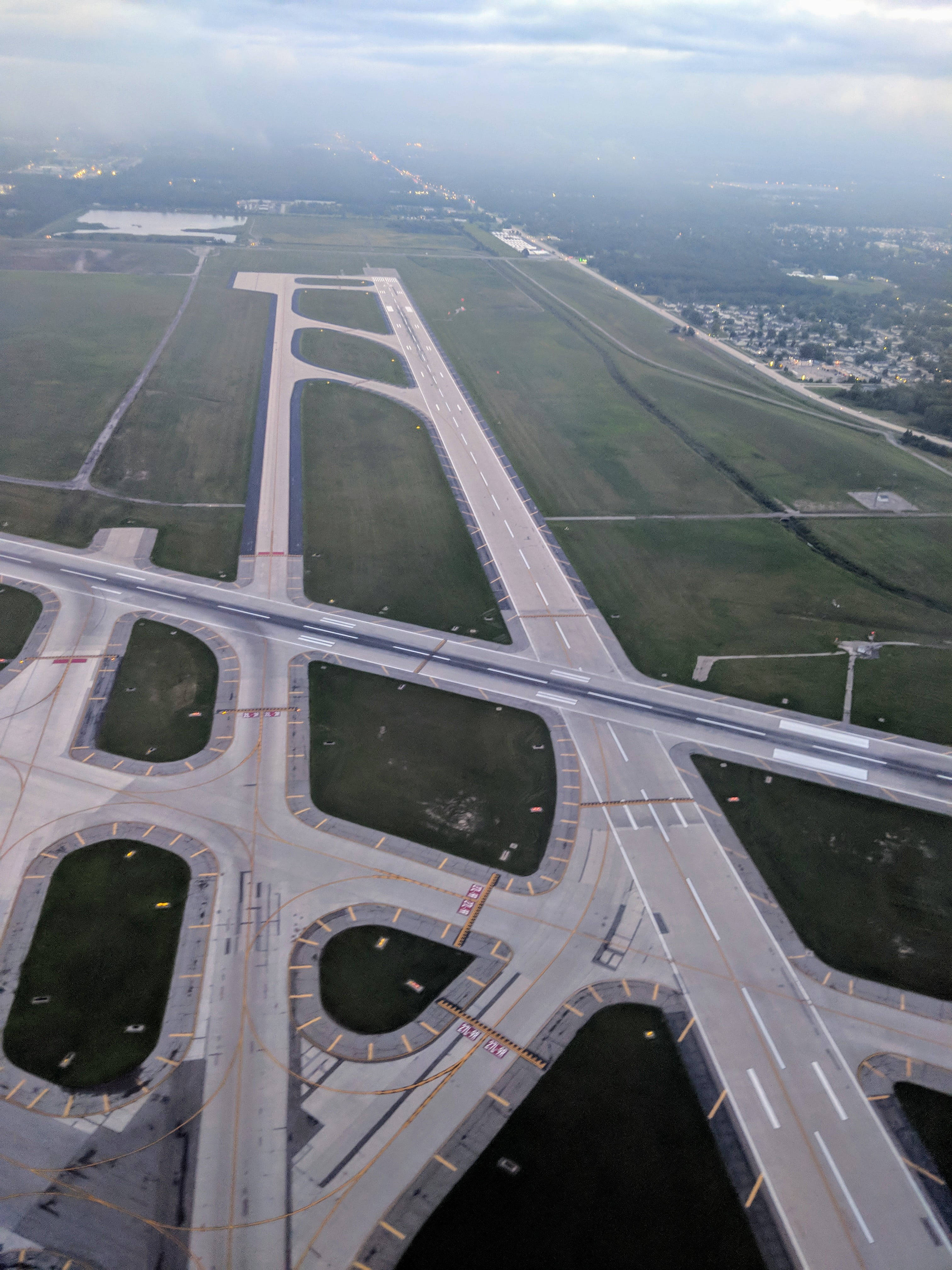 跑道空中俯瞰 壁紙 — 鳥瞰圖顯示一條廣闊的機場跑道和滑行道，與道路和綠色田野交錯，天空多雲。