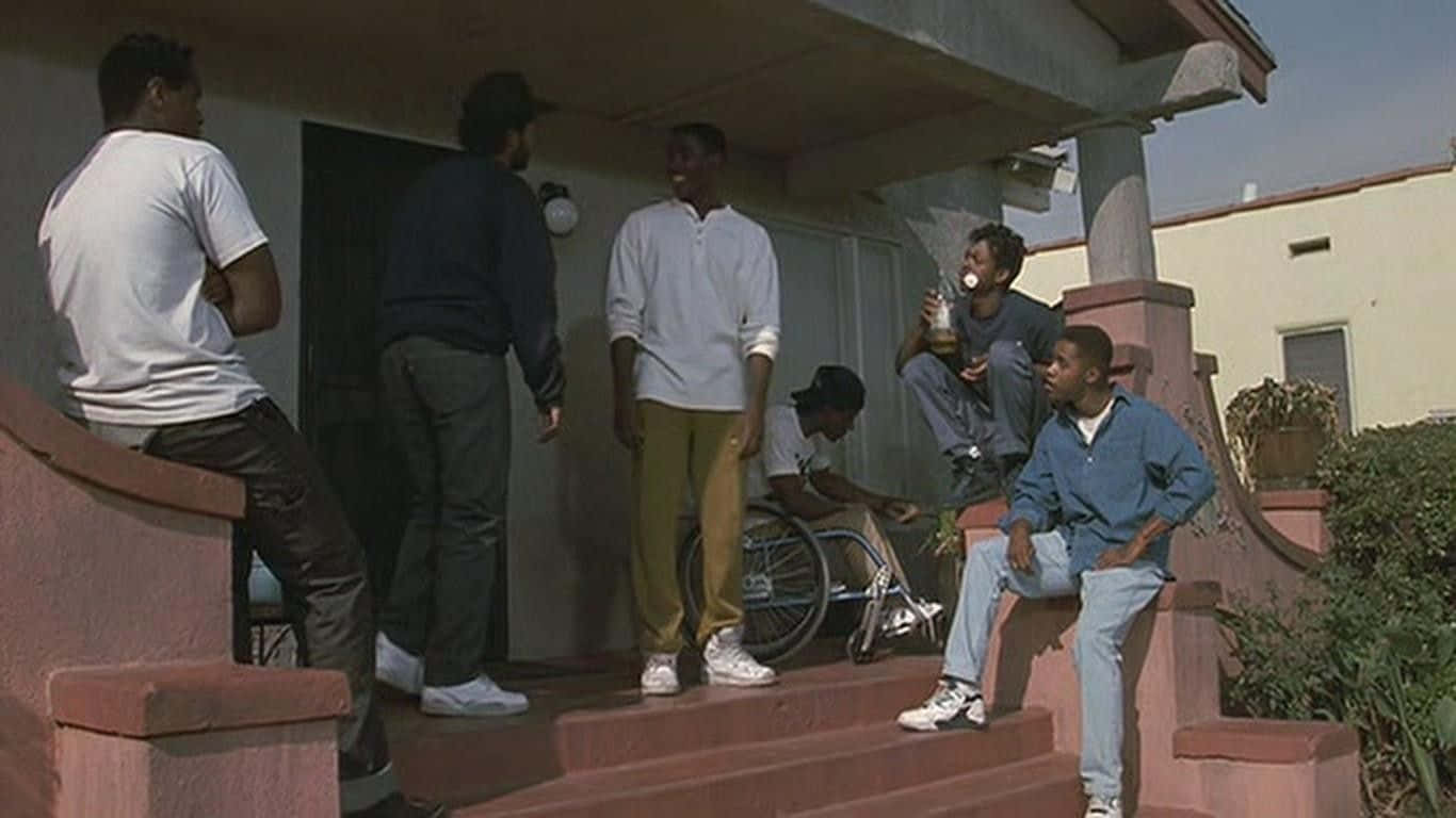 跟隨1991年經典劇情片《街区男孩》中三位童年好友的故事 壁紙 — 一群年輕男子在明亮的日光下聚集在房屋門廊的粉紅色混凝土台階上。