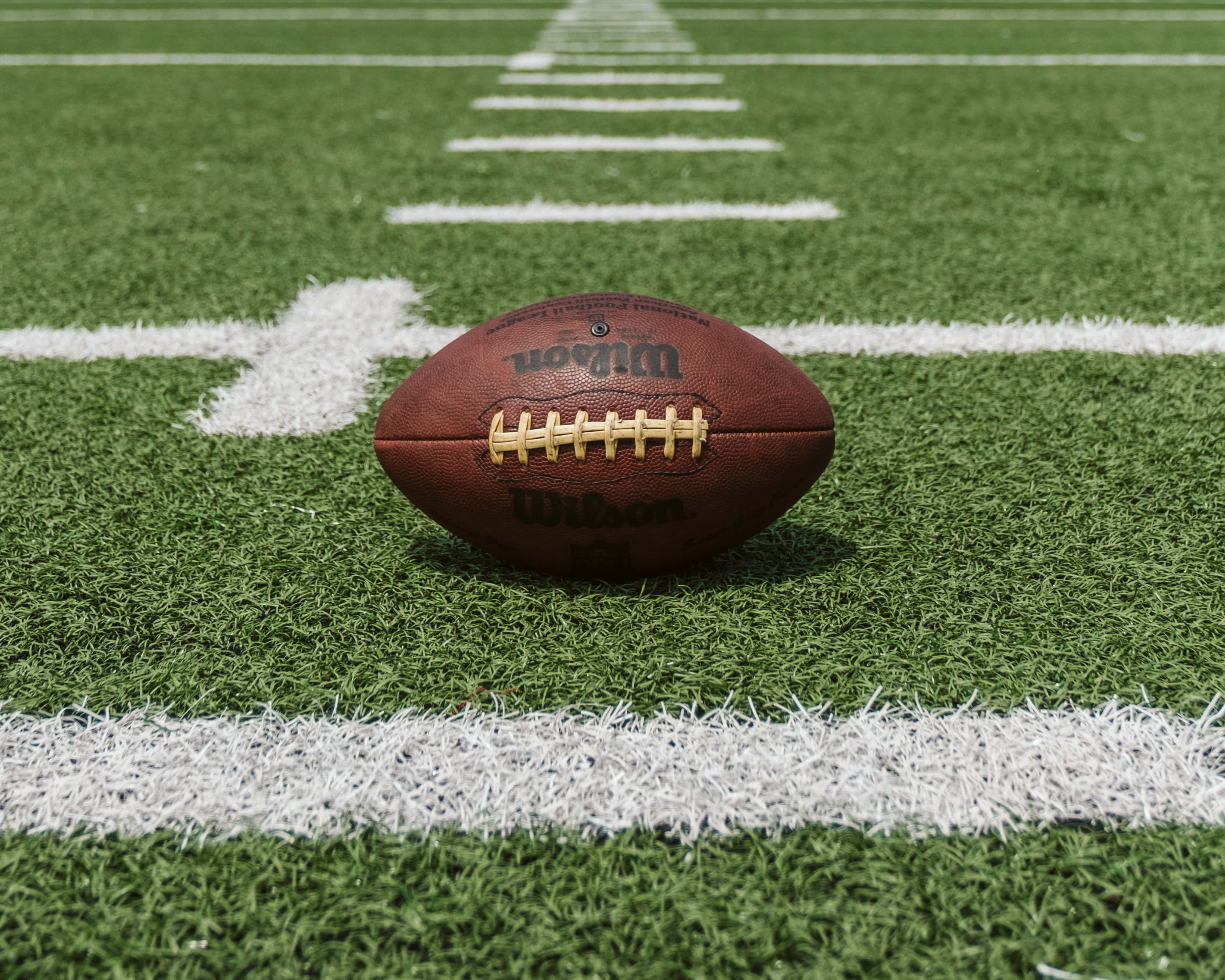 跟隨您最喜愛的NFL球隊走過2020賽季 壁紙 — 一個棕色Wilson美式足球在明亮的日光下，放置在充滿活力的綠色草地上，草地上有清晰的白色碼線。