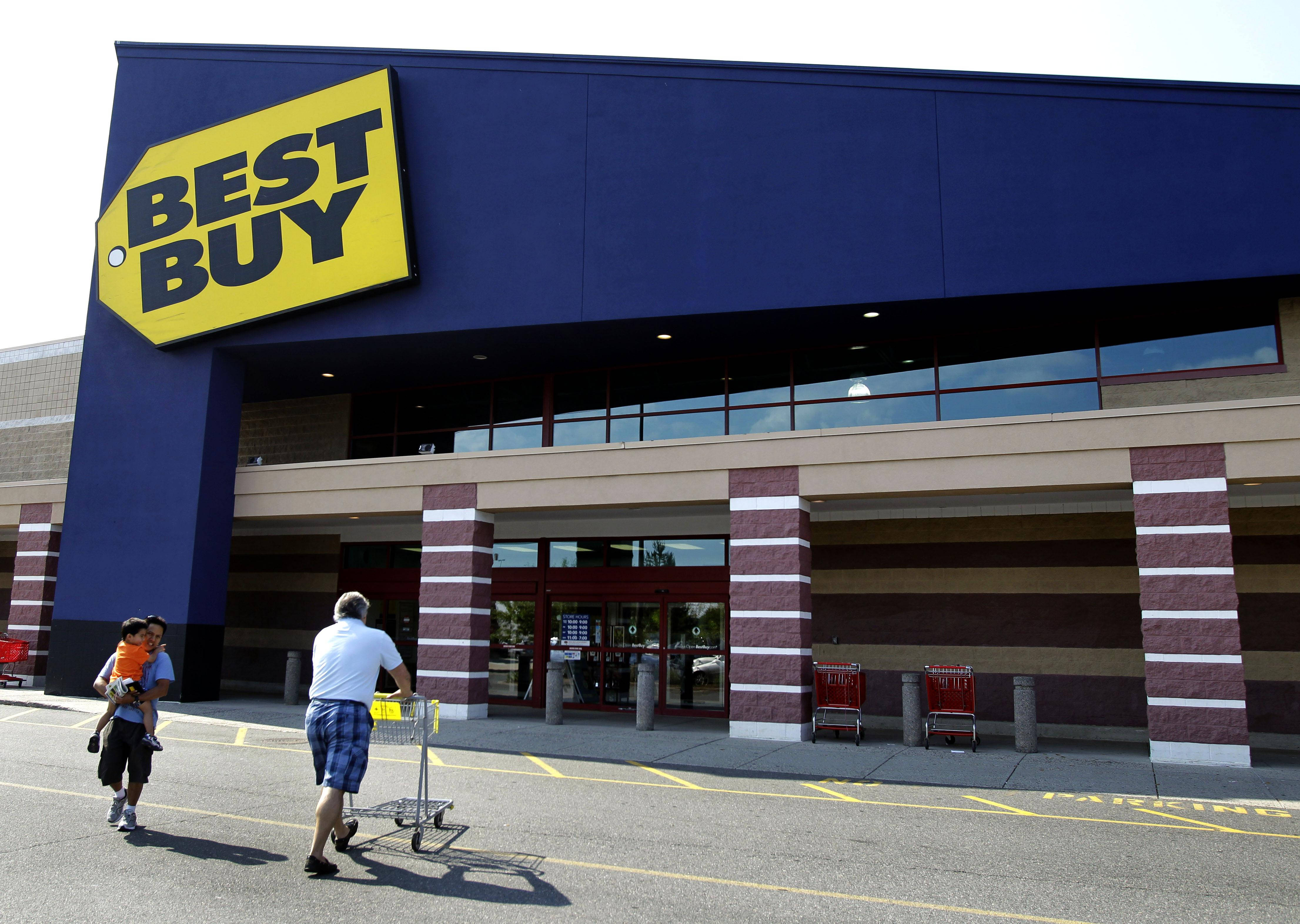 路對面的Best Buy 壁紙 — 一名男子在明亮的日光下推著購物車，同時抱著一個孩子，走向Best Buy商店的入口。