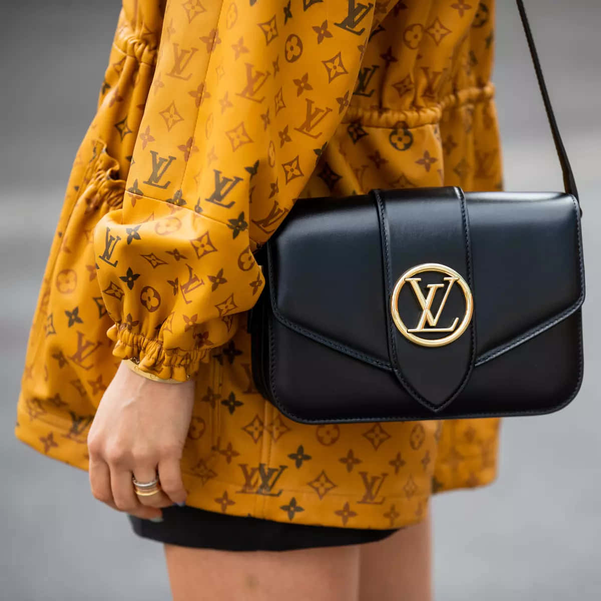 路易威登的永恆奢華 壁紙 — 近距離拍攝一個人穿著芥末黃色 Louis Vuitton 字母組合外套，並攜帶一個帶有金色標誌的黑色 LV 手袋。