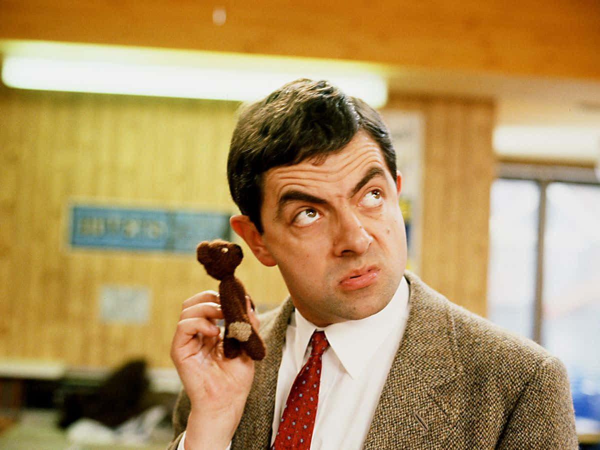 Mr Bean 桌布 壁紙 — 路雲·雅堅遜飾演Mr Bean，在辦公室裡抱著一隻細小的棕色泰迪熊，露出滑稽而懷疑的表情。