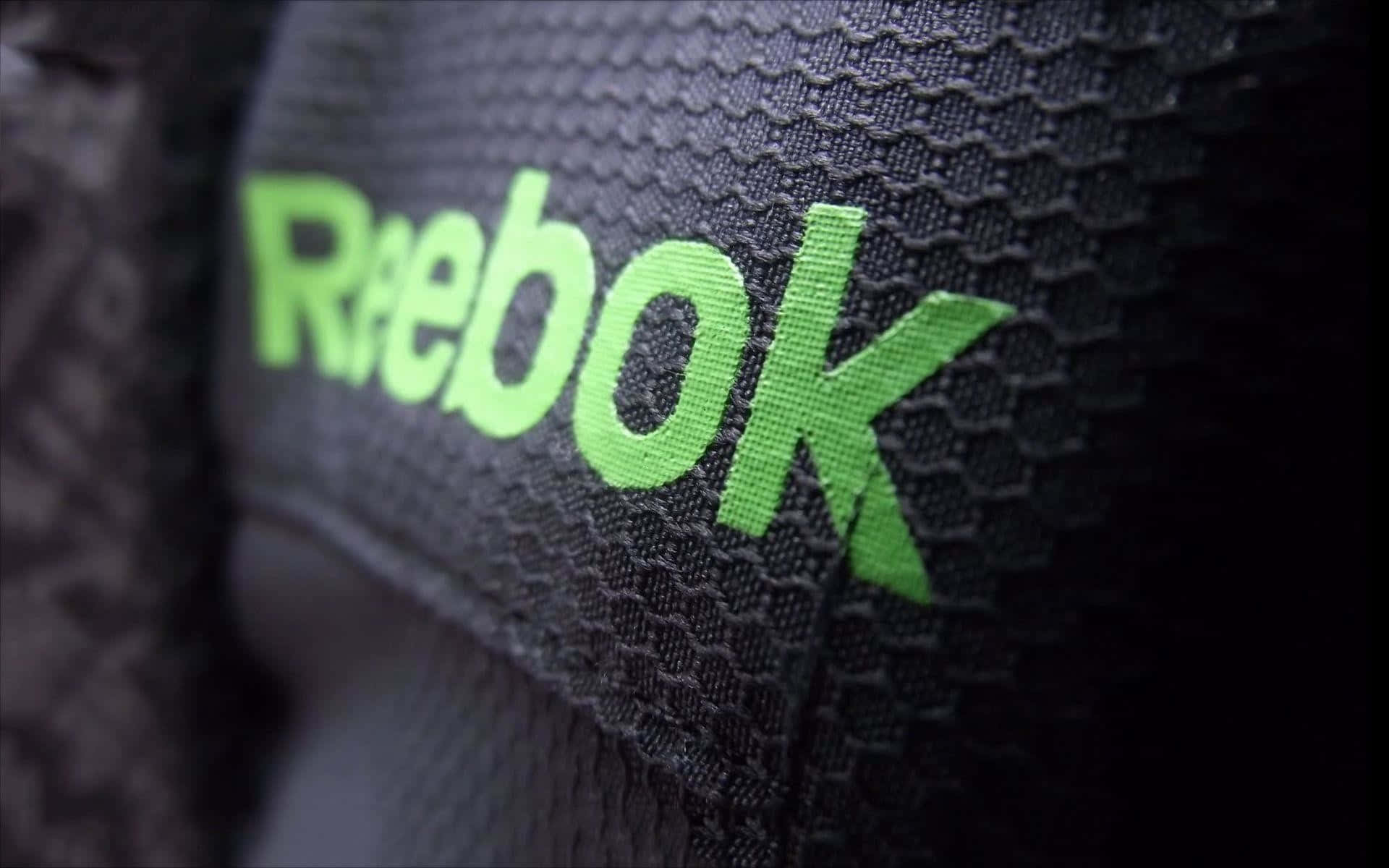 踏入Reebok，準備迎接世界挑戰 壁紙 — 鮮綠色Reebok標誌近距離特寫，縫在帶有紋理的黑色運動布料上，景深較淺。