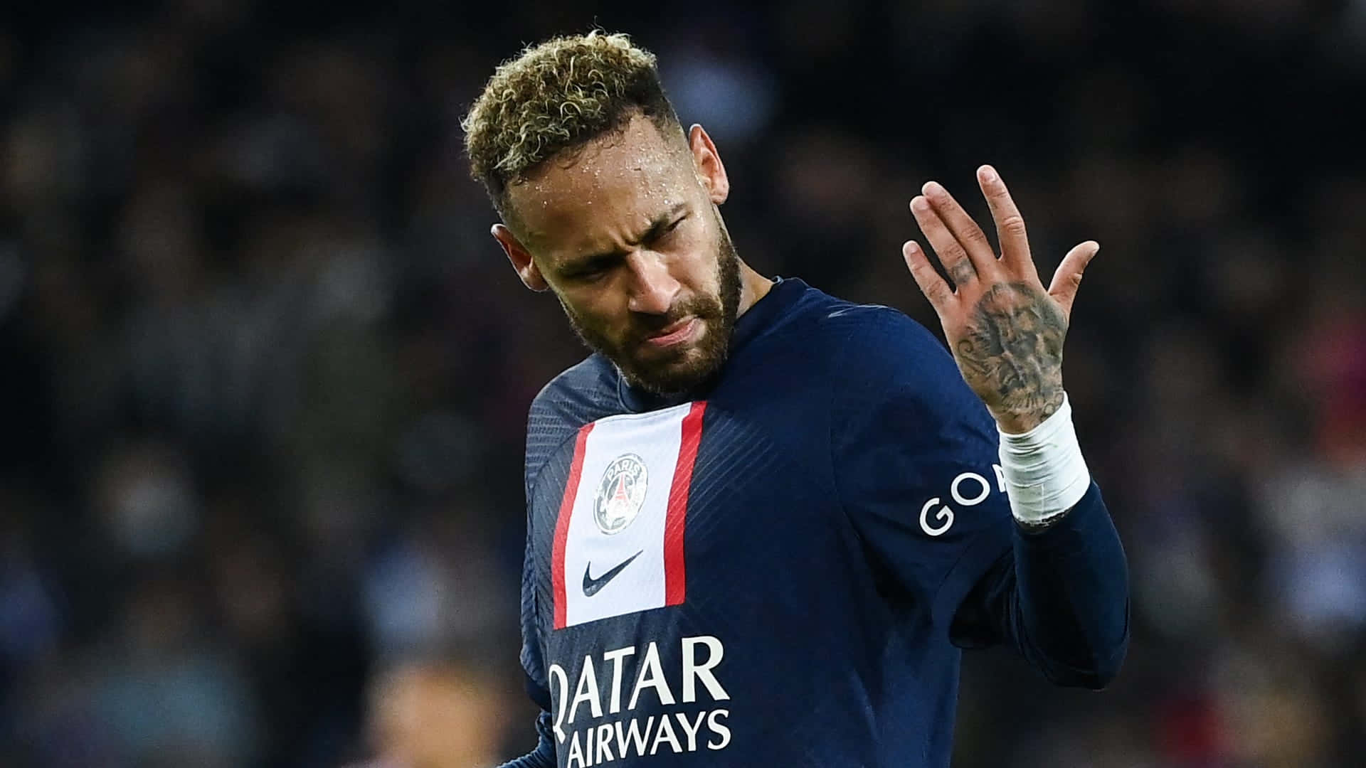 Neymar 桌布 壁紙 — 身穿PSG球衣的足球員尼馬在夜間比賽中，在球場燈光下用有紋身的手向人群揮手。