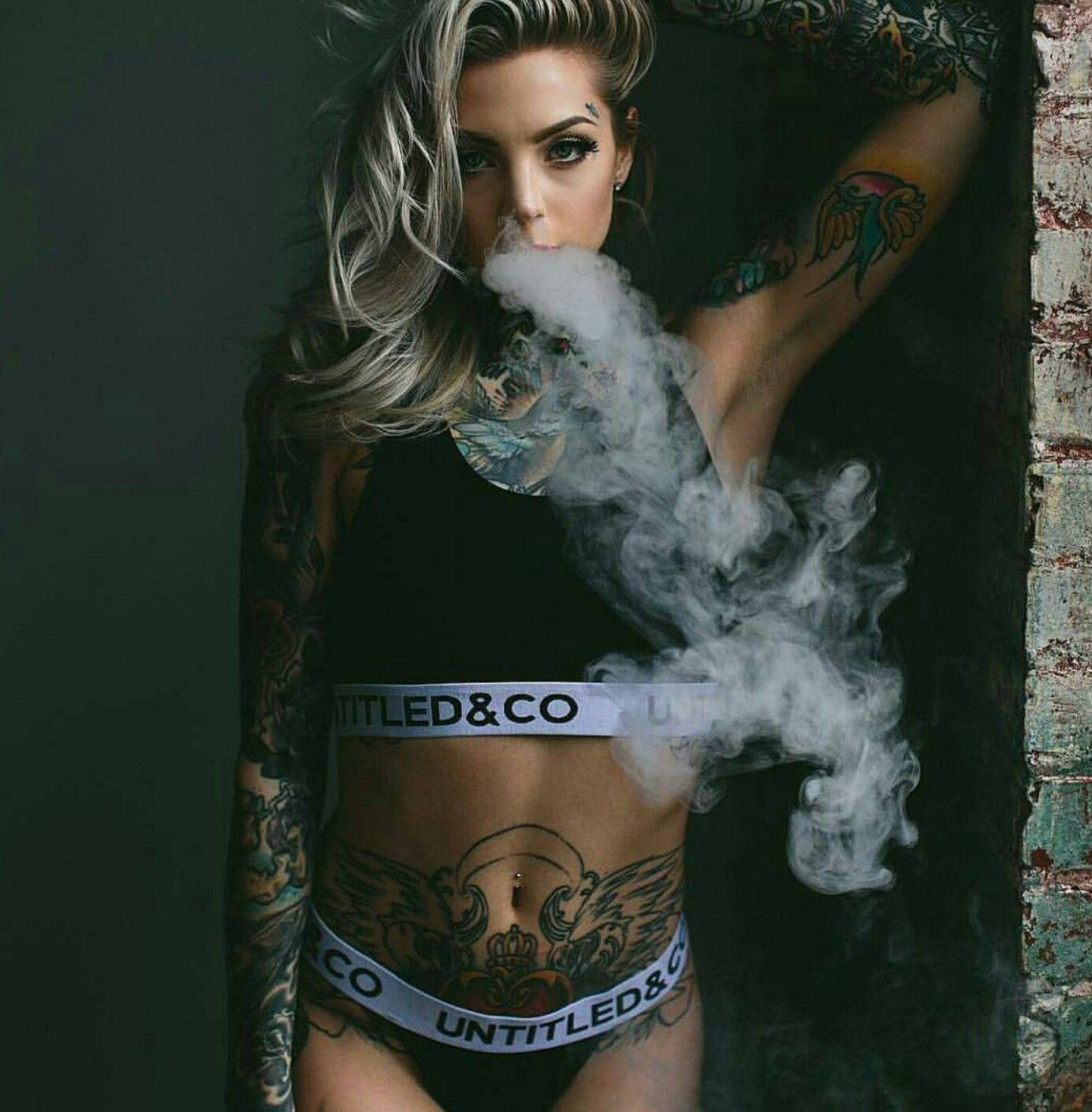 身穿Untitled & Co. 品牌胸衣的性感美女模特 壁紙 — 一位金髮紋身女性在深色紋理牆壁前穿著品牌內衣呼出煙霧。