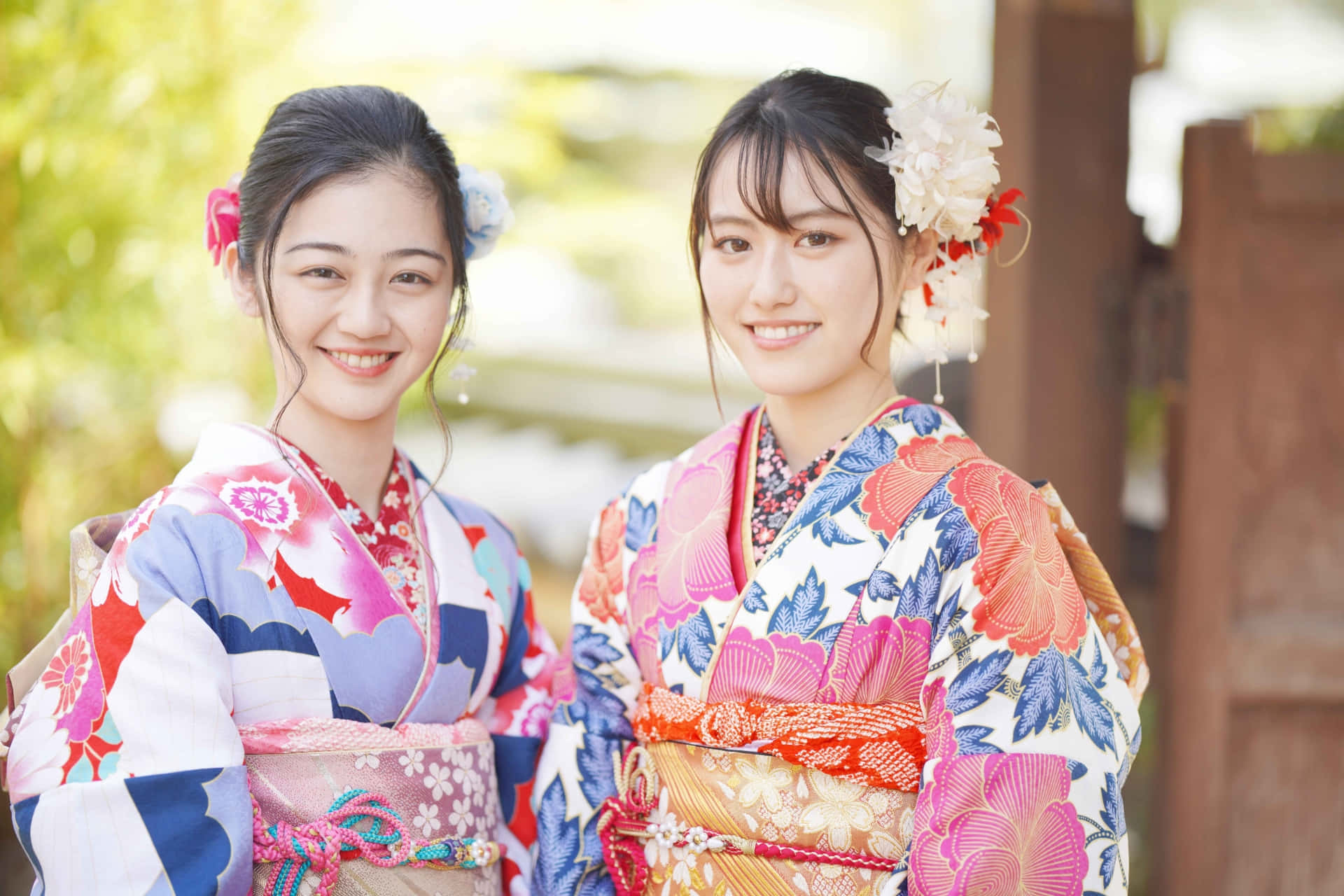 身穿傳統日本和服的女性 壁紙 — 兩位微笑的年輕女性穿著鮮豔的花卉和服，在戶外擺姿勢，柔和的光線透過她們身後的綠植灑落下來。