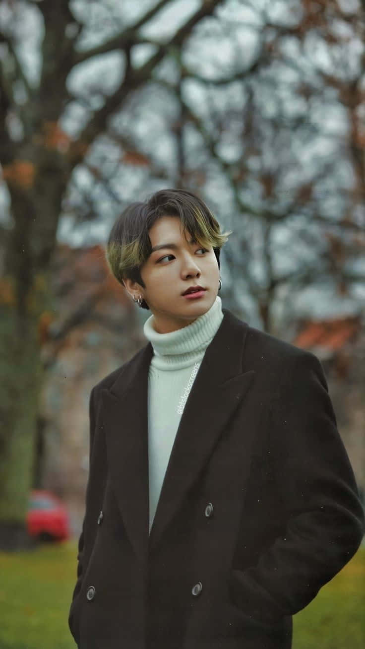 身穿時尚黑色服裝的神秘Jungkook 壁紙 — 一位年輕男子，頭髮造型時尚，戴著耳環，身穿黑色外套內搭白色高領毛衣，站在公園裡，背景是光禿的樹木和柔和的秋色。